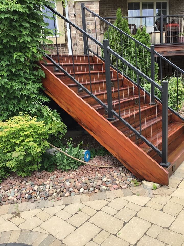Wooden outdoor staircase with black railings beside a house and garden shrubs