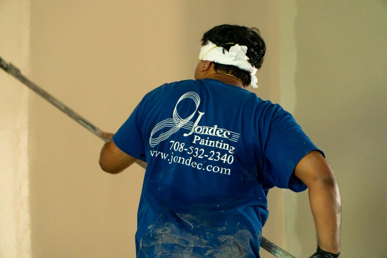 Painter in blue shirt applying paint to a wall with a roller, seen from behind.