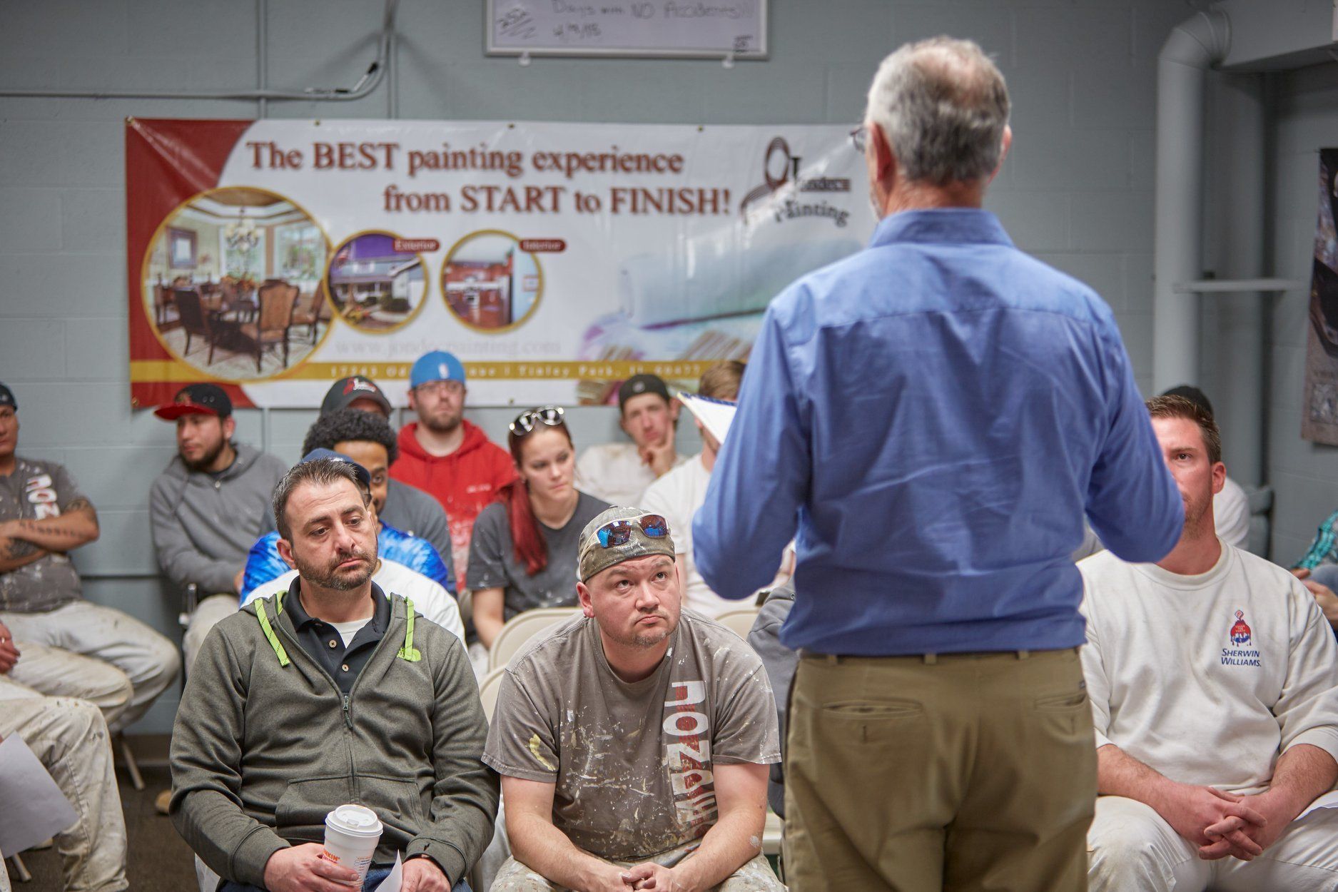Man speaking to a seated group in a room with a painter services banner