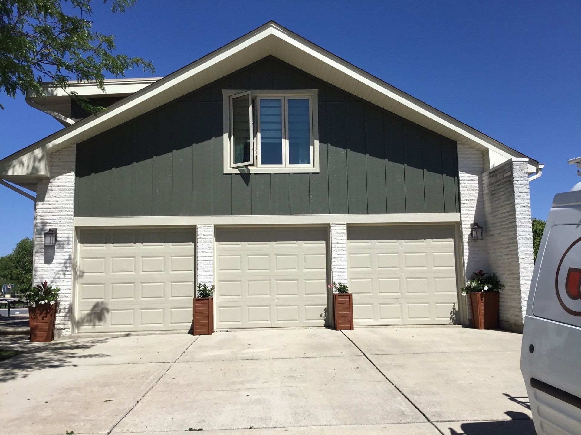 Suburban house with three garage doors, gray siding, and a white parked vehicle at the right edge.