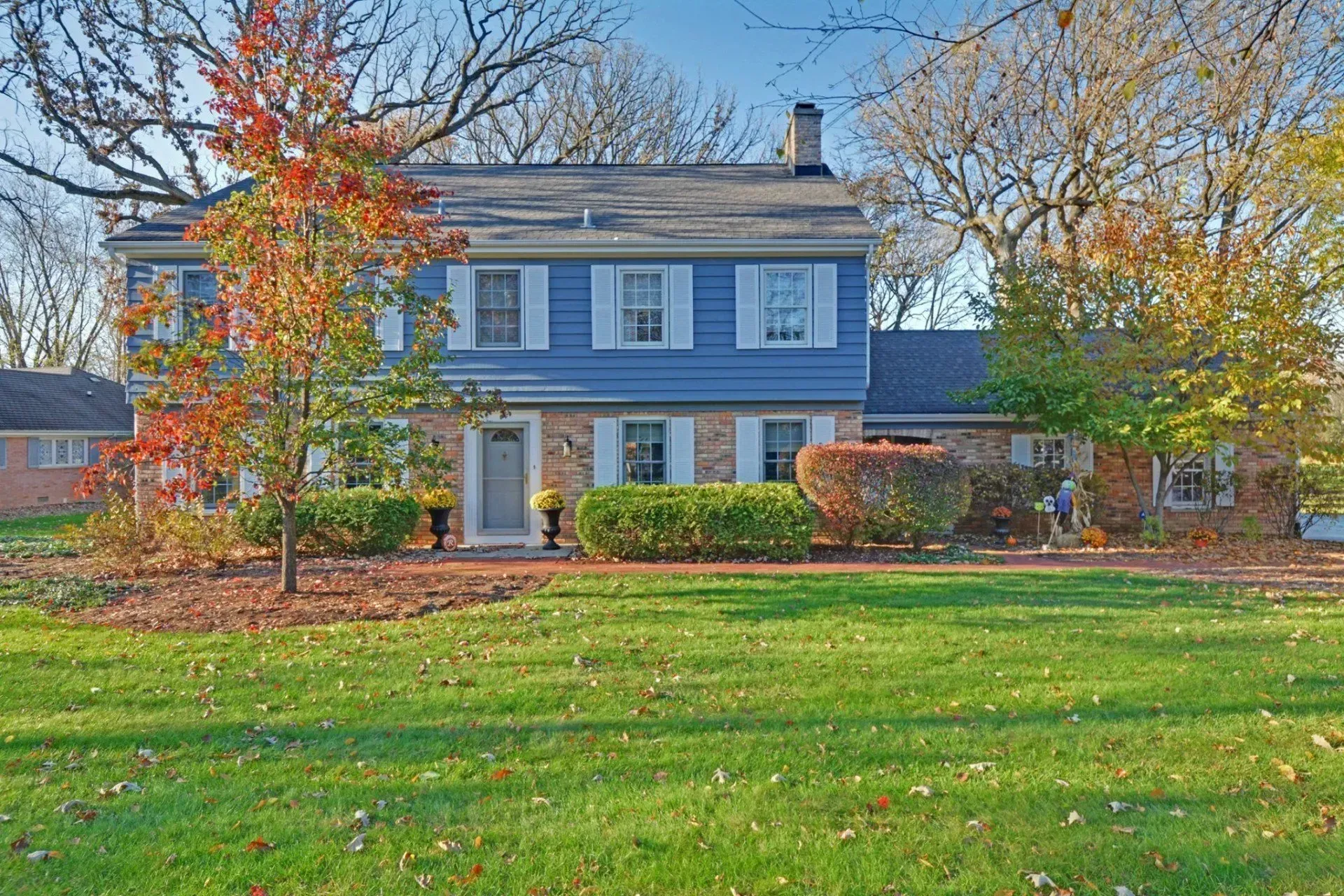 Two-story blue house with white trim and a large green lawn in autumn, framed by colorful trees.