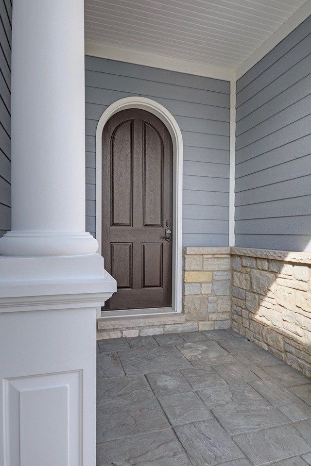 Front porch with arched brown door, white columns, gray siding, and stone wall trim