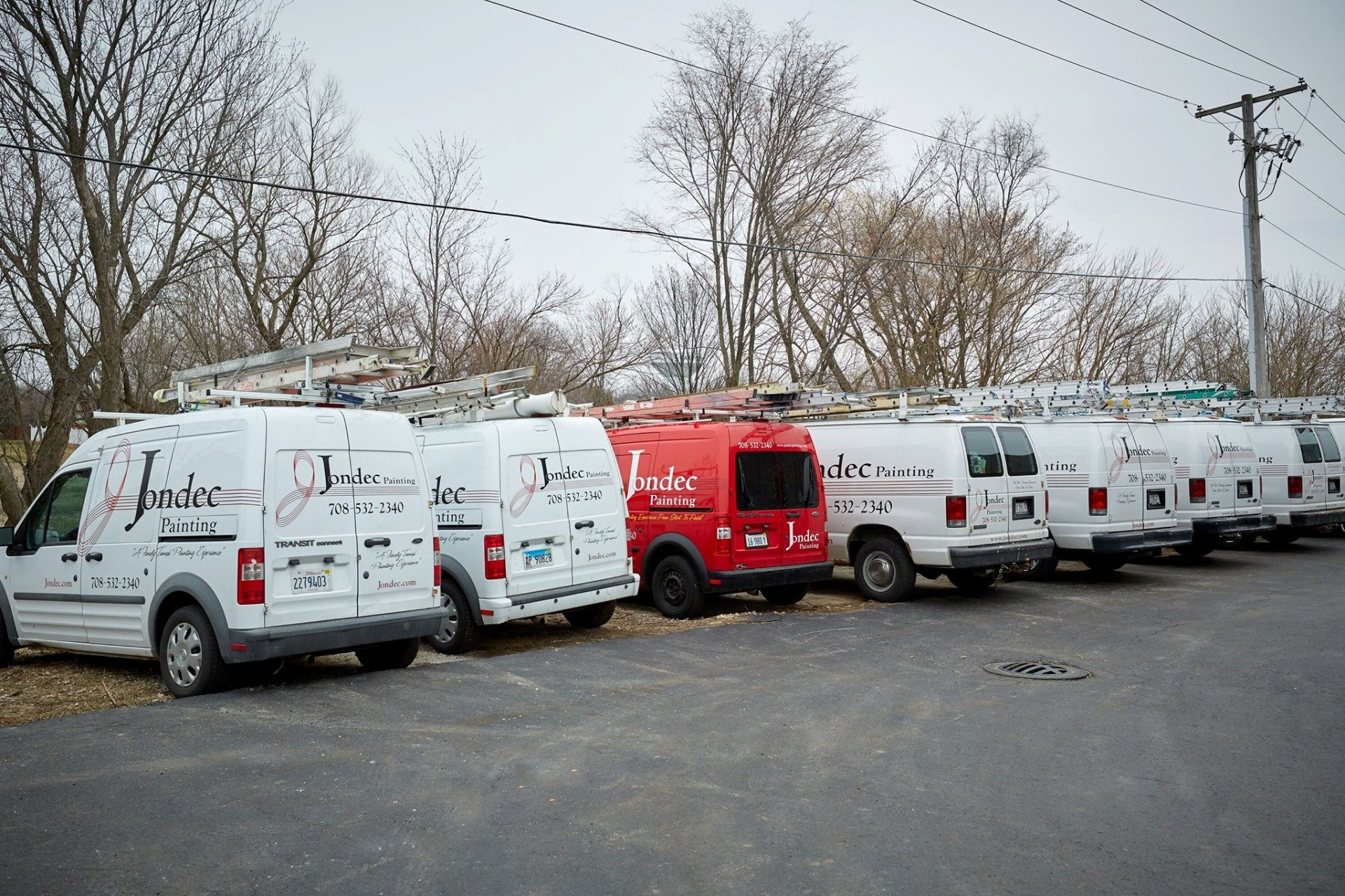 Line of white and one red utility vans parked outdoors on a gravel lot under overcast sky