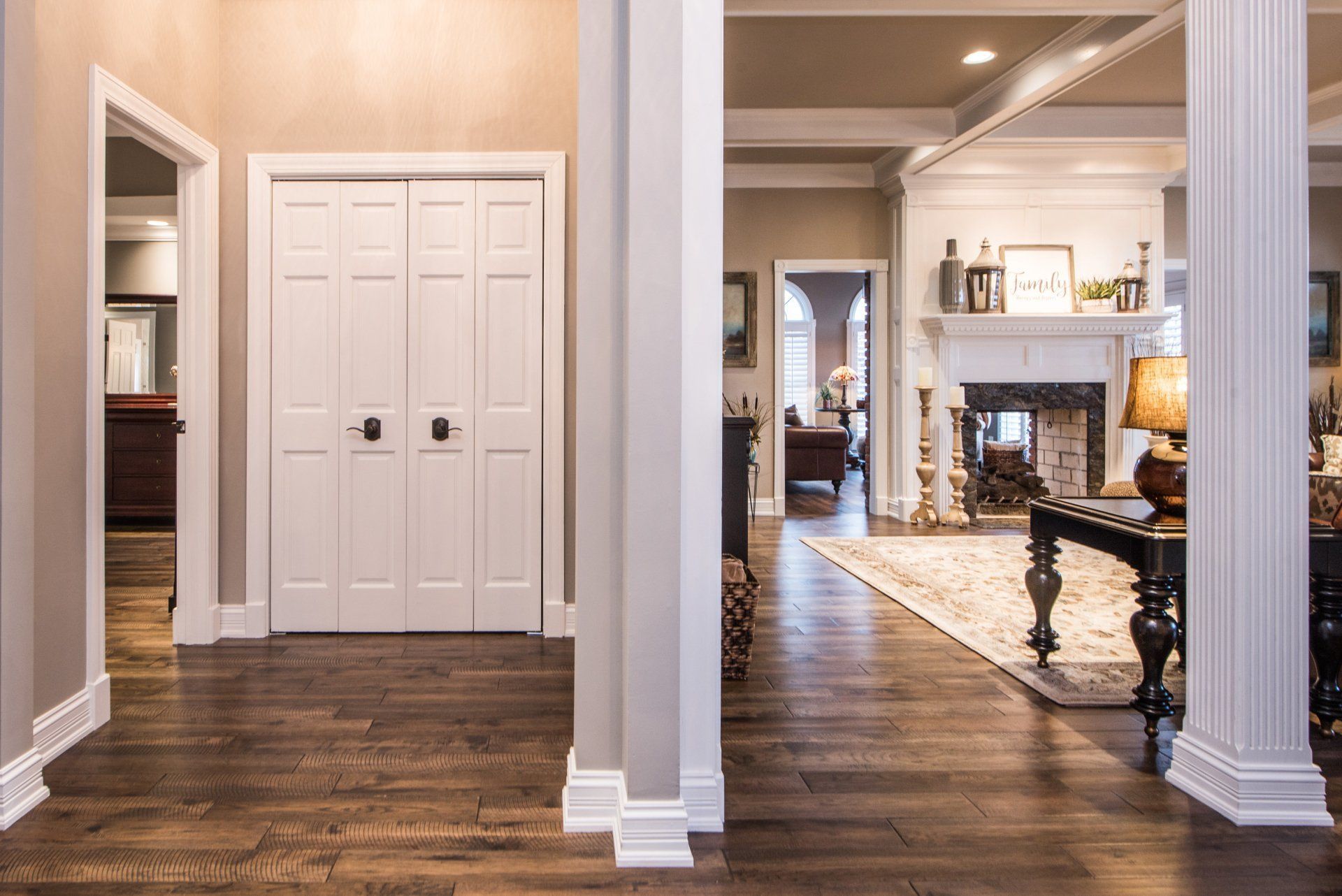 Bright foyer with dark wood floors, white columns, and a hallway opening into a living room with a fireplace.