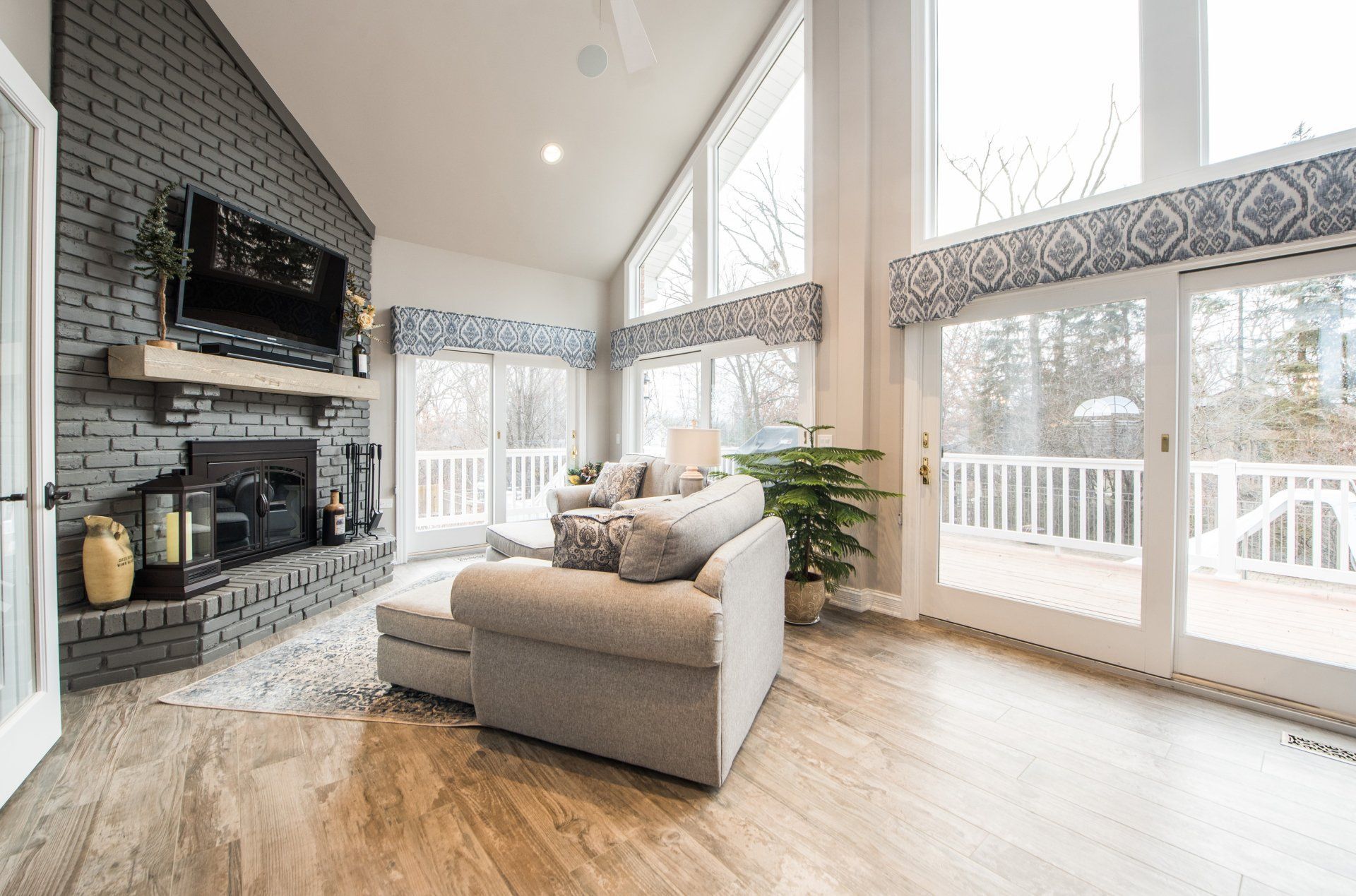 Bright living room with a stone fireplace, beige sofa, and large windows overlooking a deck.