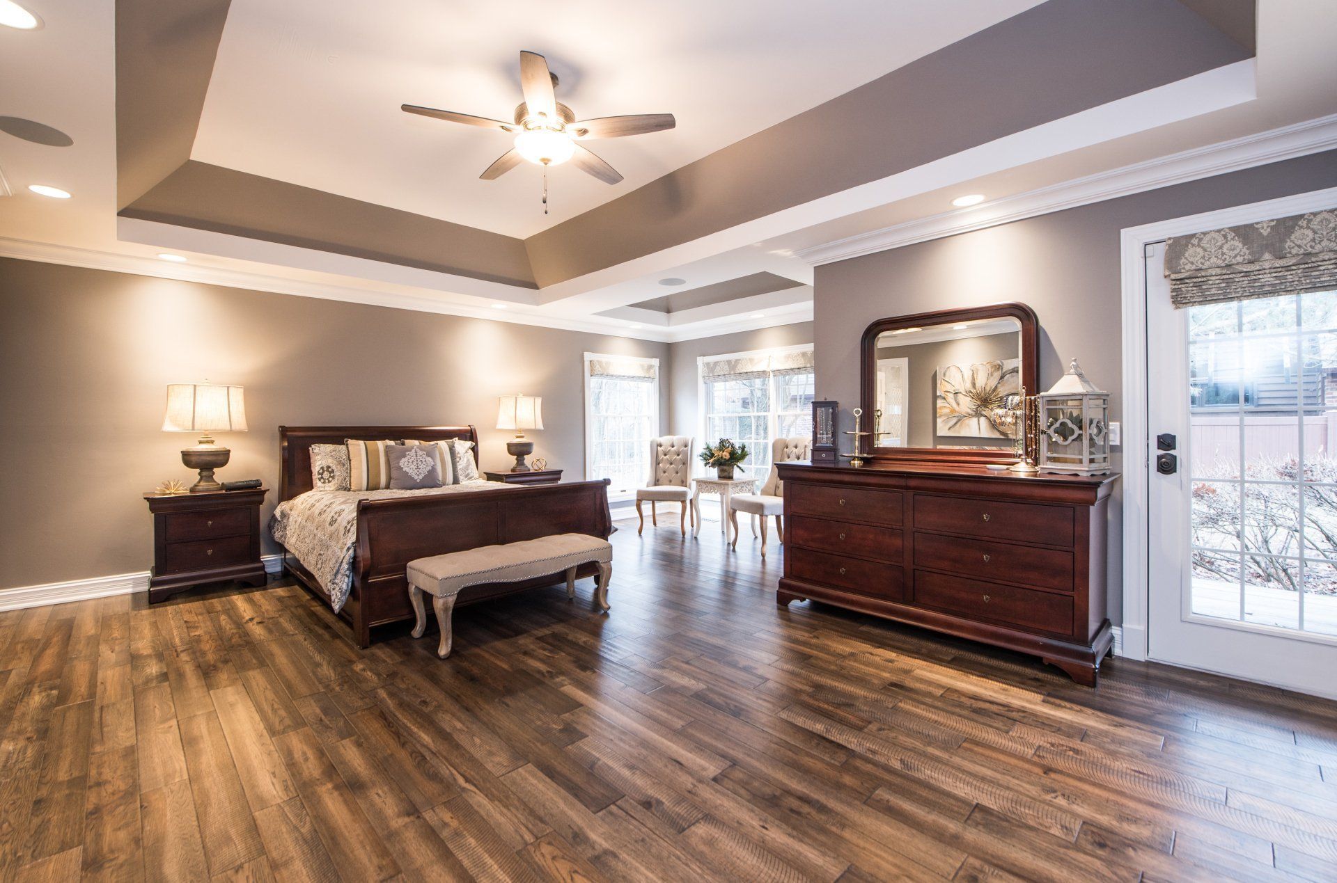 Spacious bedroom with wood floors, dark wood furniture, and a large bed under a tray ceiling with ceiling fan.
