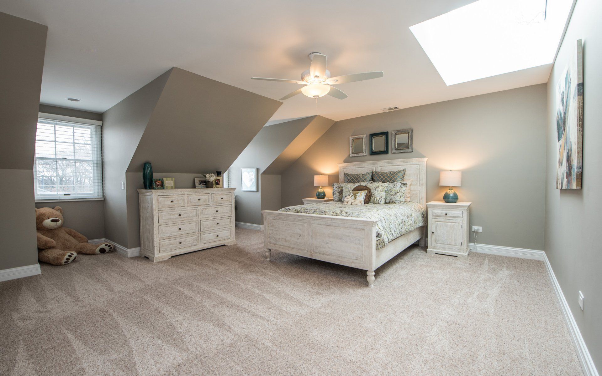 Spacious beige bedroom with a bed, dresser, teddy bear, skylight, and sloped ceiling
