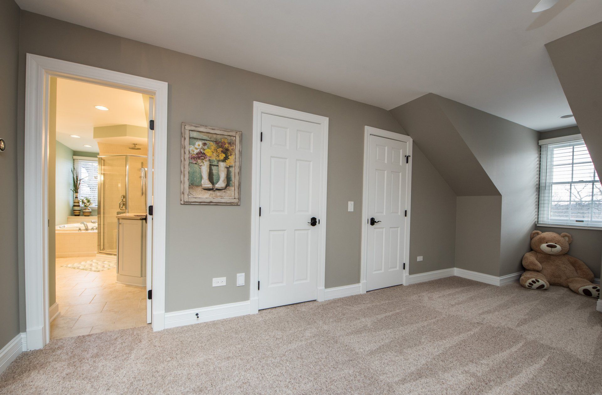 Carpeted beige bedroom with white double closets, open doorway to a bathroom, and teddy bear by the window.