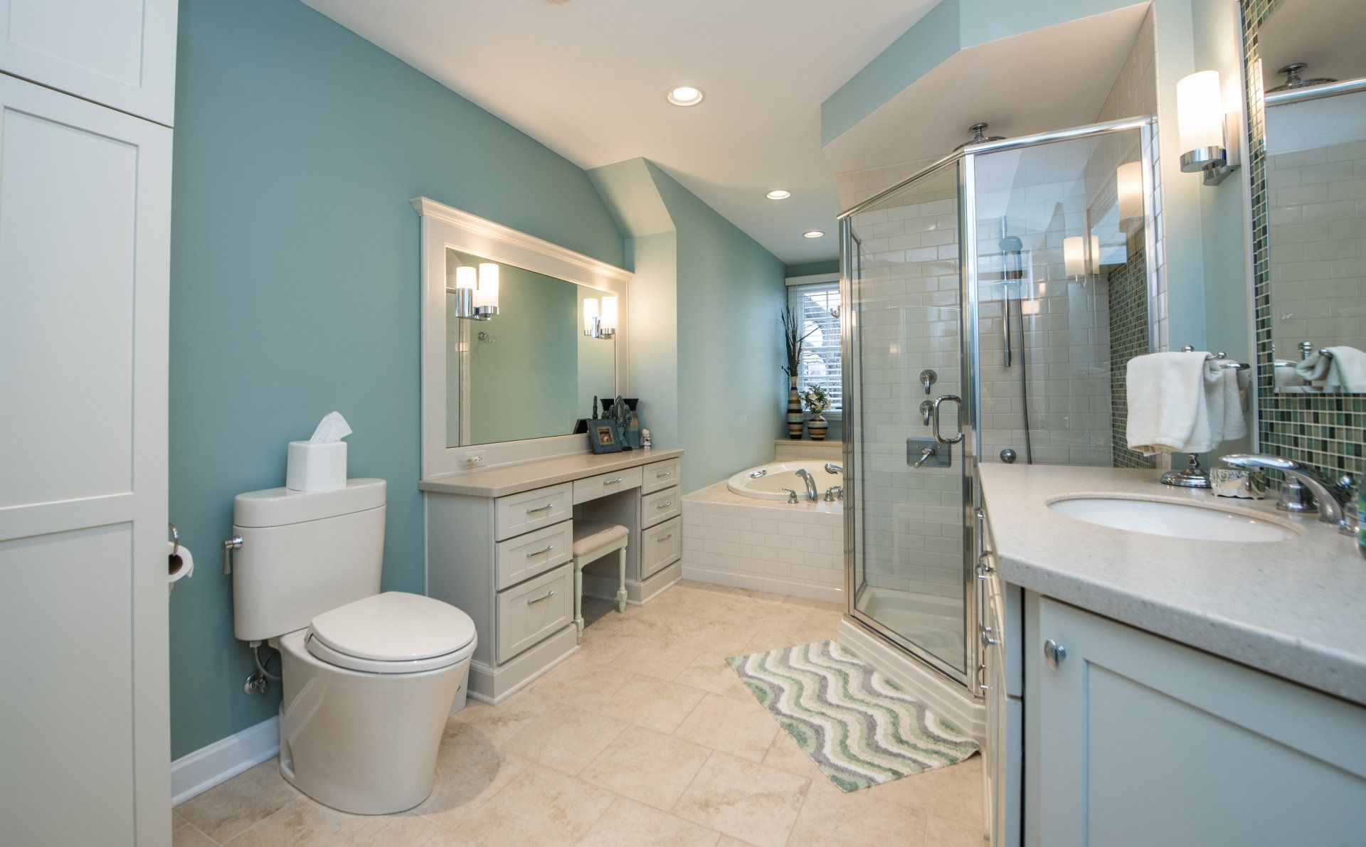 Bright modern bathroom with dual vanity, toilet, soaking tub, glass shower, and light blue walls.