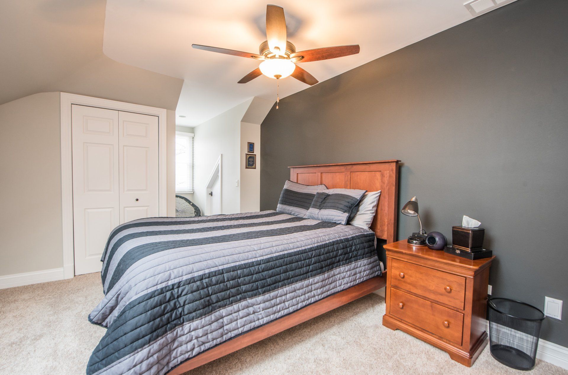 Bedroom with a neatly made bed, gray accent wall, white closet doors, and a wooden nightstand with lamp.