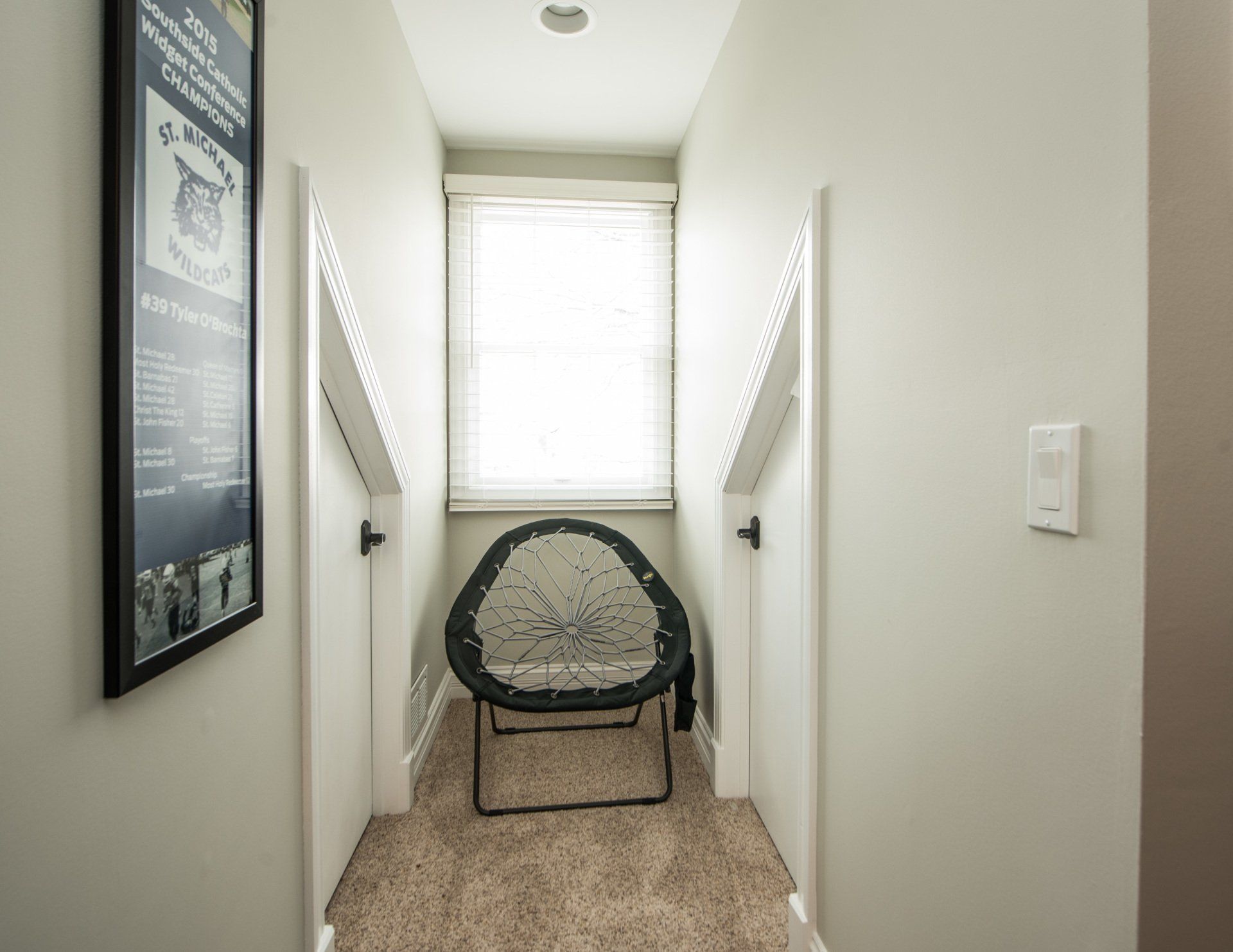 Narrow hallway with a woven chair under a window and white walls, carpeted floor, and handrails on both sides