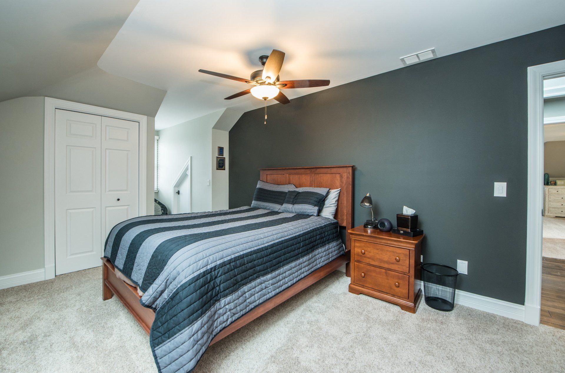 Bedroom with a wooden bed, gray accent wall, ceiling fan, and nightstand on carpeted floor