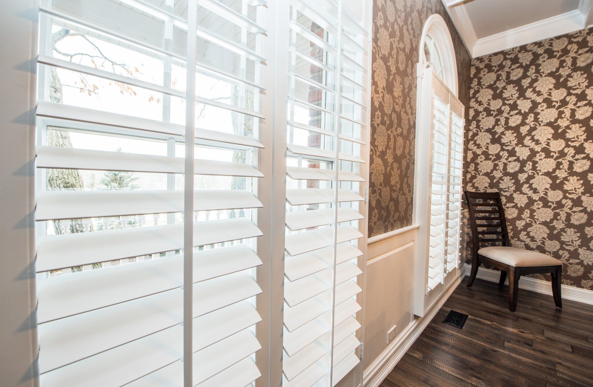 Bright sunroom with white plantation shutters, patterned wallpaper, dark wood floor, and a chair by a small table.