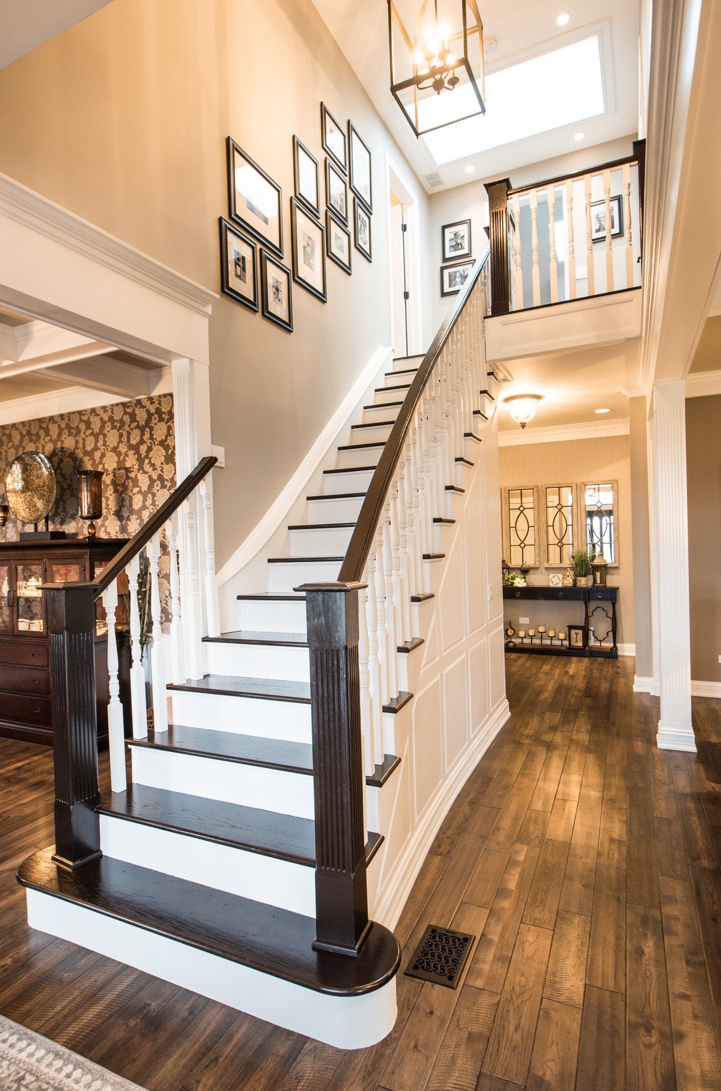 Bright foyer with a white staircase, dark wood floors, framed wall art, and warm lighting.