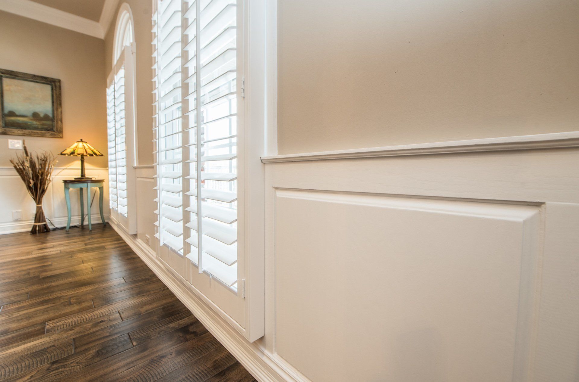 Bright hallway with white shutters, wood floor, and a small table with lamp and decor.