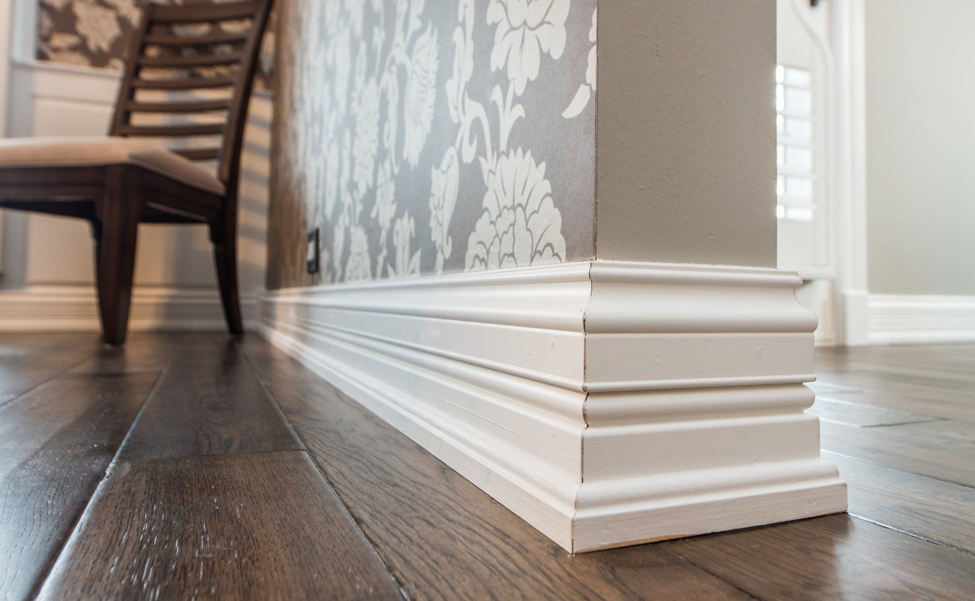 White decorative baseboard trim on a hardwood floor beside patterned wallpaper and a chair leg.