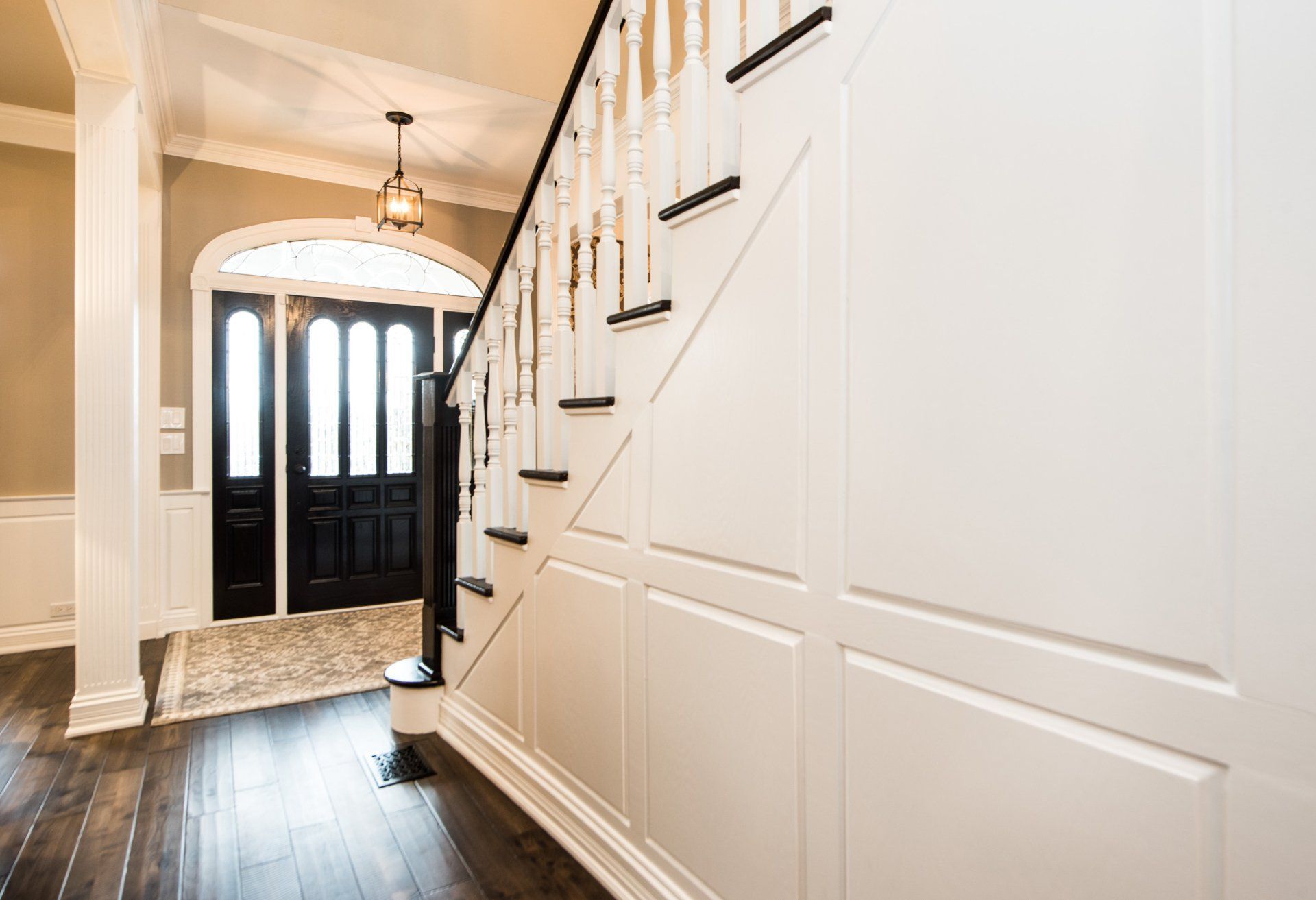 Bright hallway with white paneled walls, dark wood floor, black front door, and staircase railing.