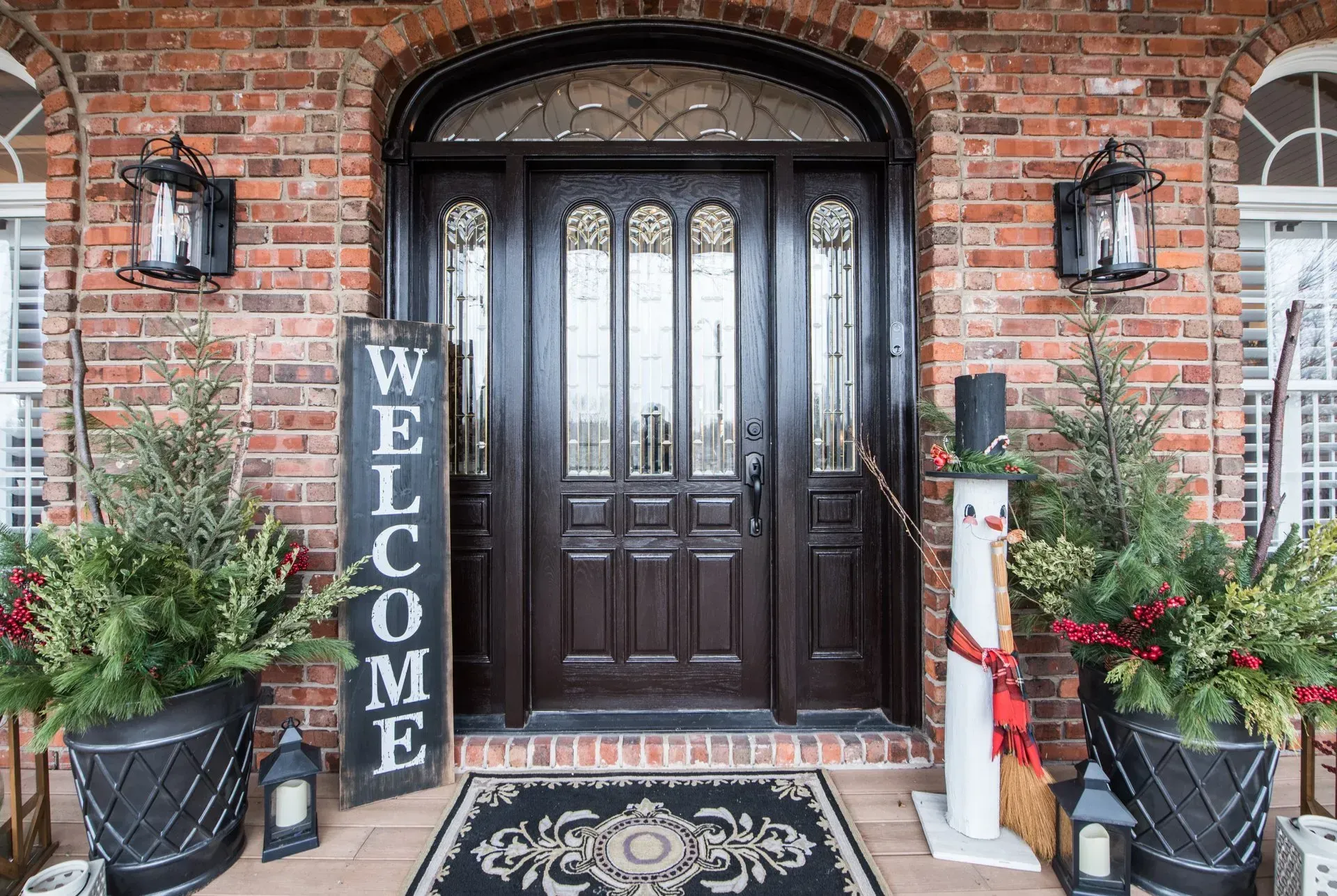 Brick home entryway with dark wood door, welcome sign, potted plants, and holiday decor on either side