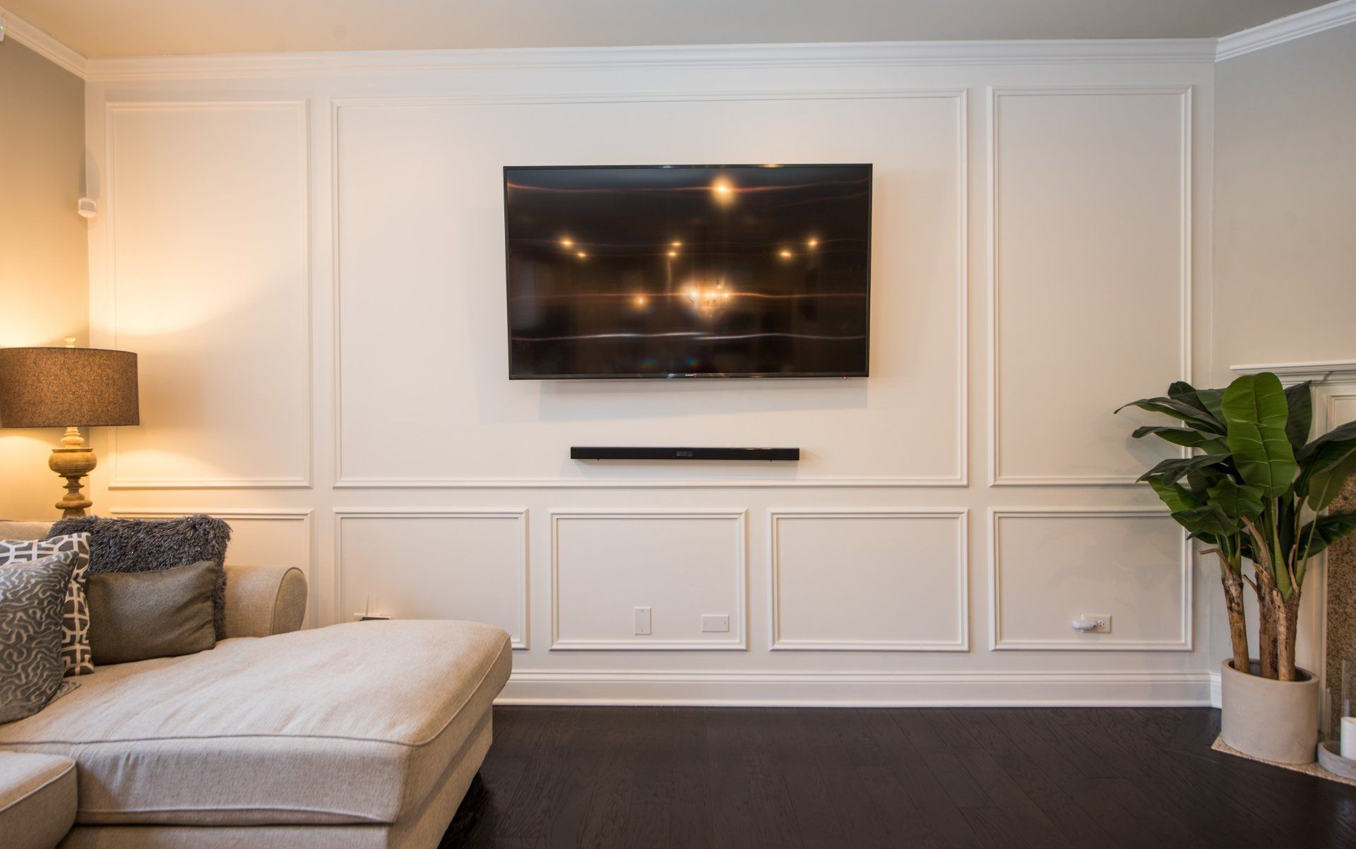 Modern living room with wall-mounted TV, beige sofa, lamp, and potted plant against white paneled walls