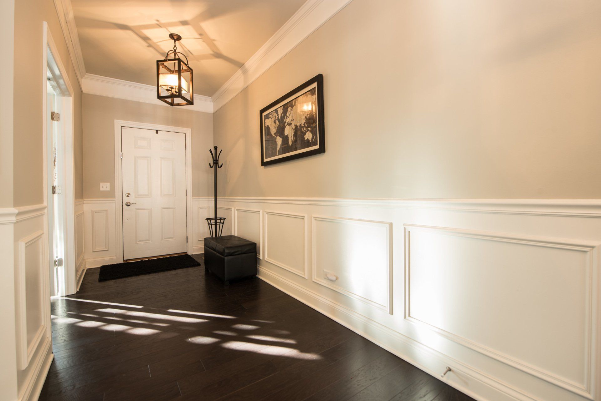 Bright hallway with white wainscoting, dark floor, front door, bench, coat rack, and hanging lantern light.