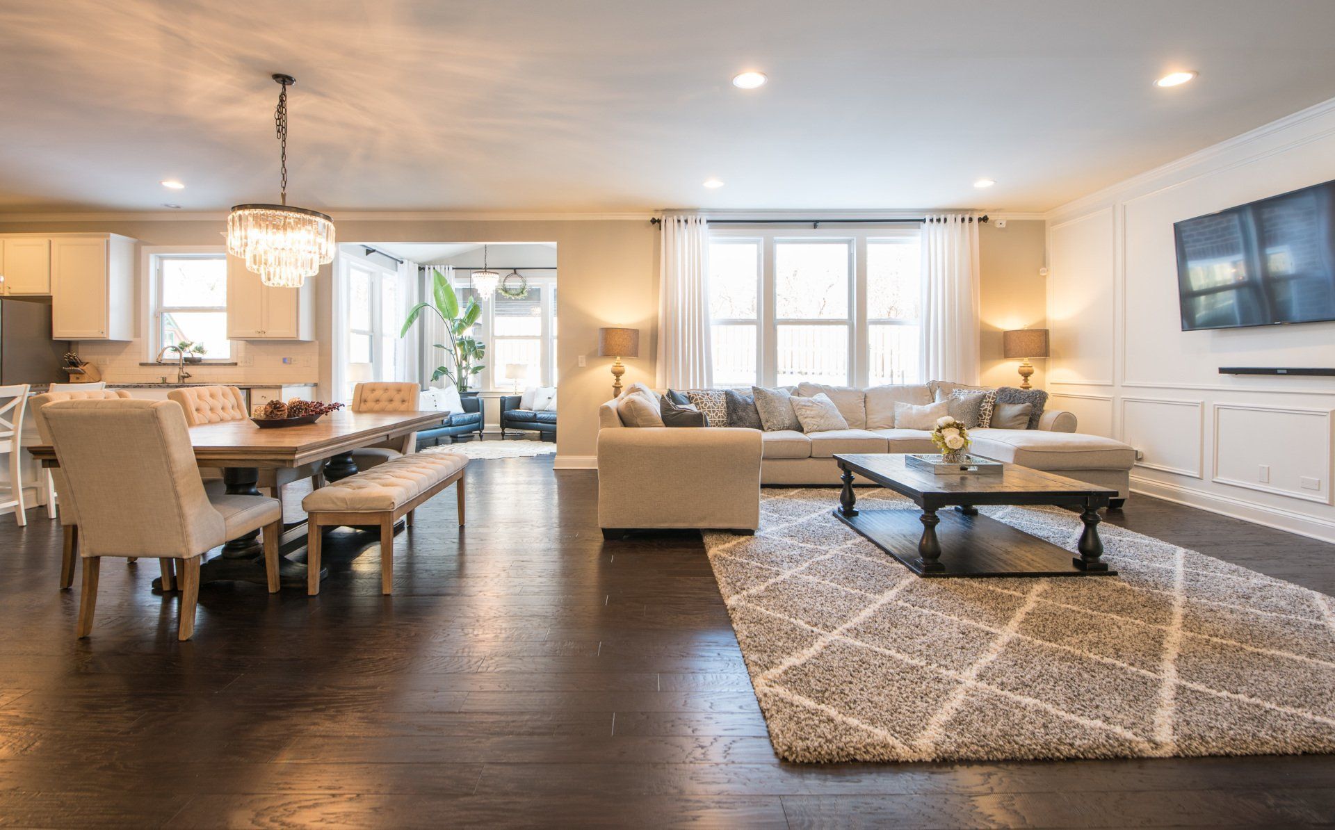 Bright open living and dining room with dark wood floors, beige furniture, chandelier, and TV on the wall