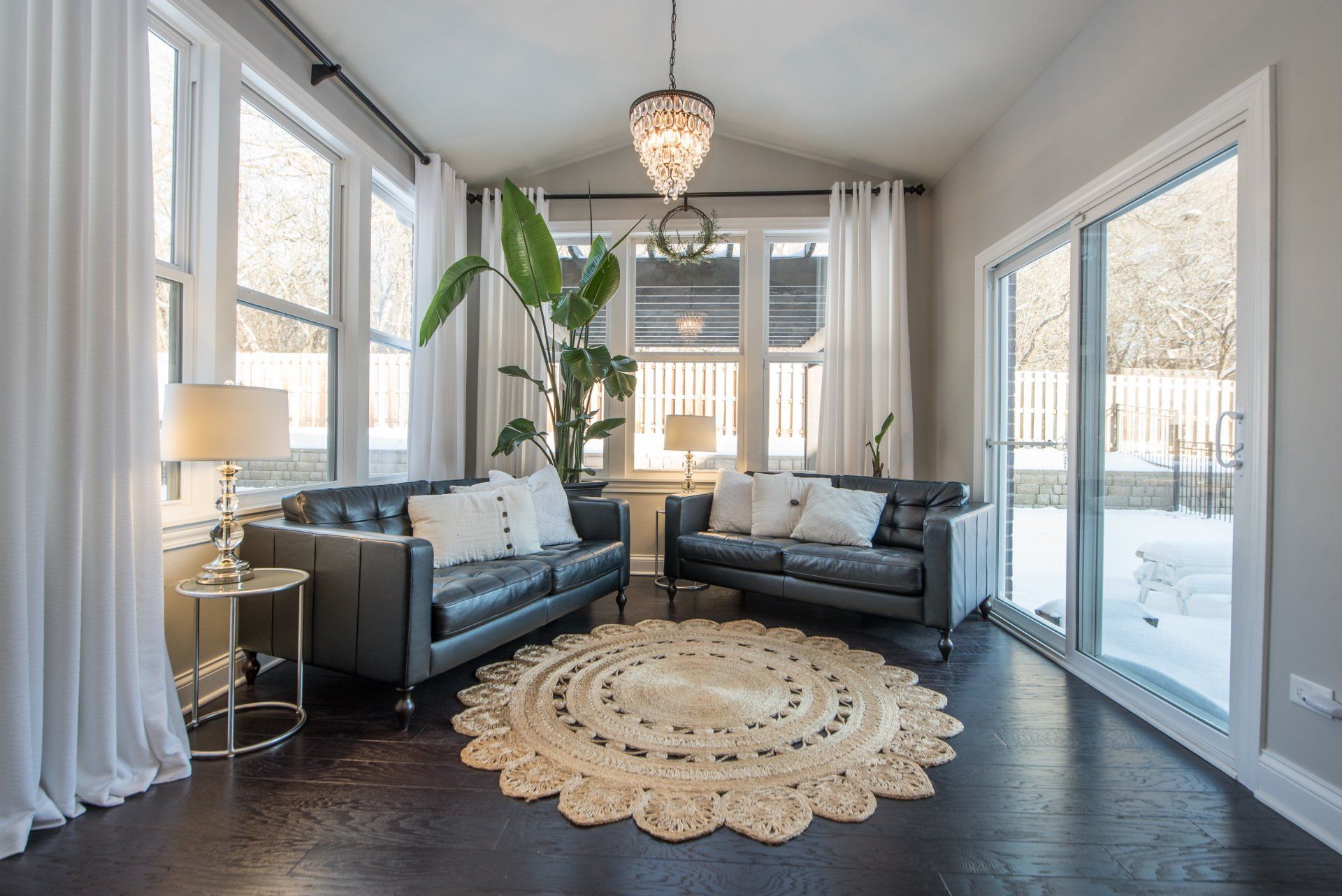 Bright sunroom with dark sofas, large windows, a round rug, and a potted plant