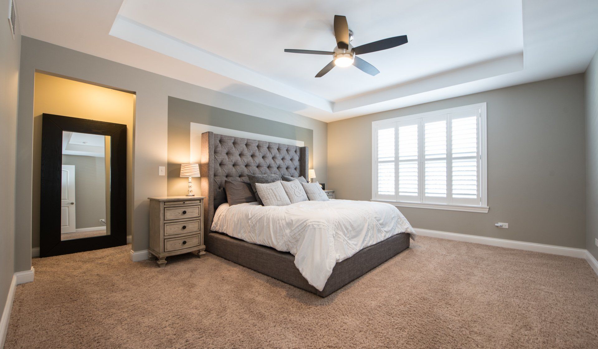 Spacious bedroom with gray tufted bed, beige carpet, ceiling fan, and bright window shutters