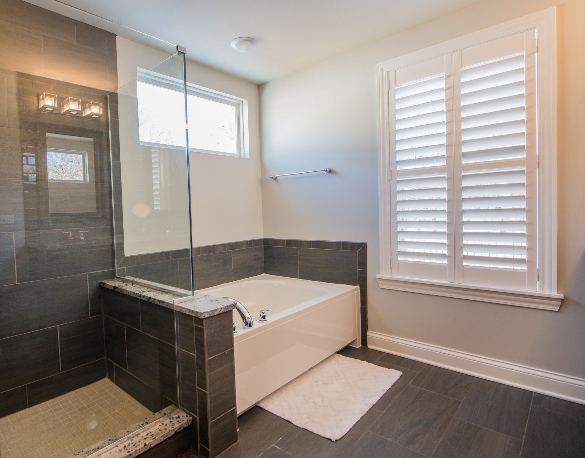 Modern bathroom with a glass-enclosed tub, dark tile, and white plantation shutters.