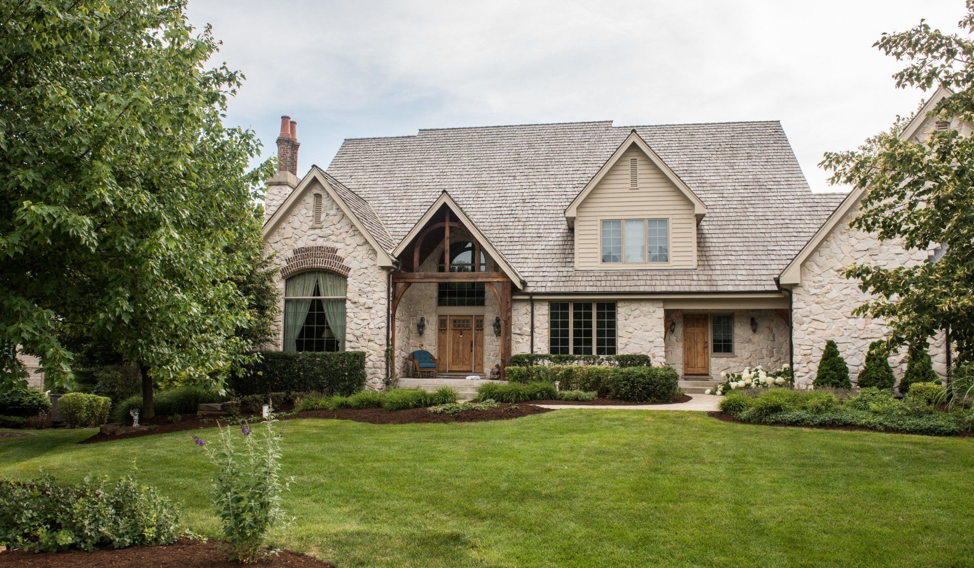Large stone suburban house with a steep roof, manicured lawn, and mature trees