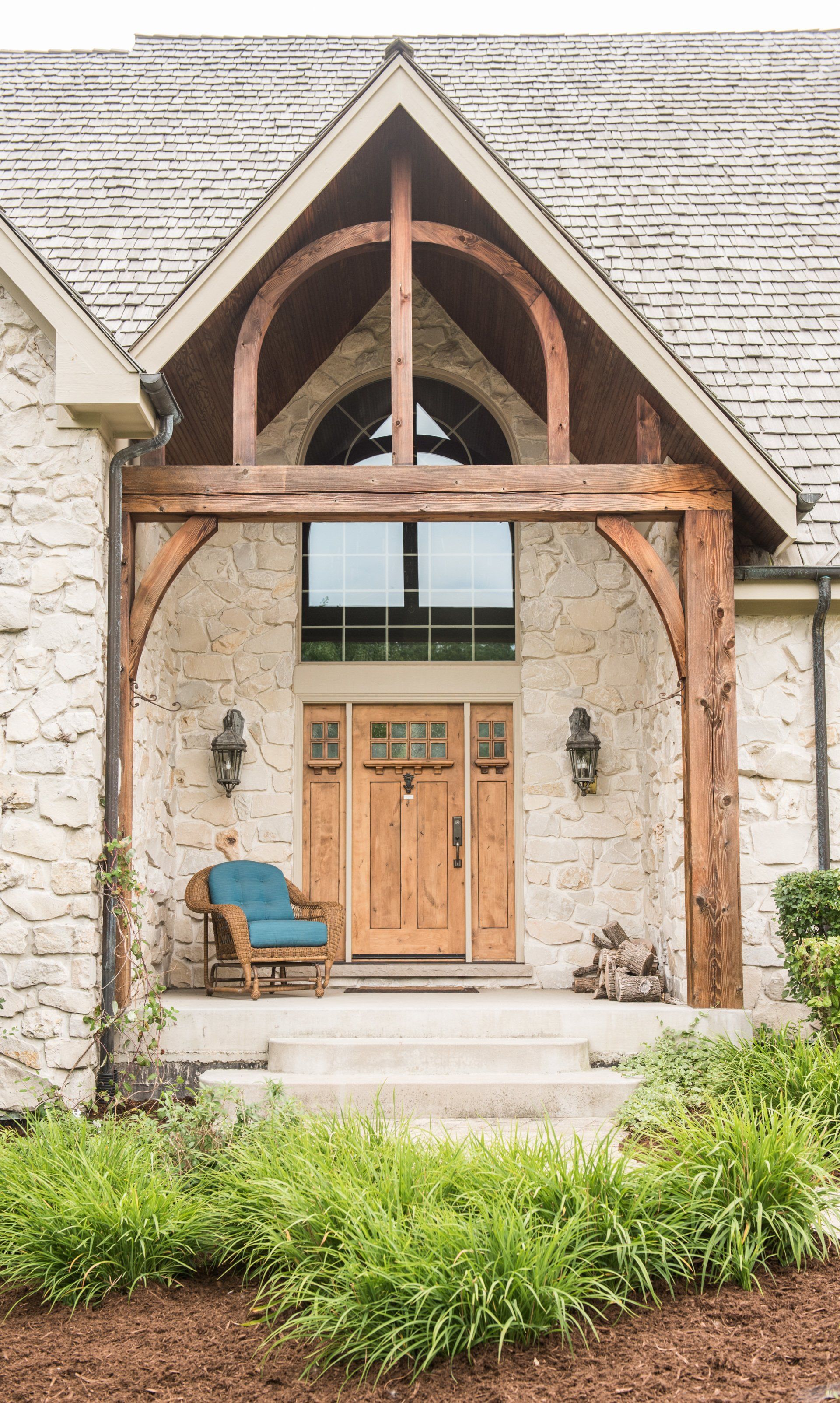 Stone house entry with wooden doors, arched timber porch, and blue chairs on a front patio