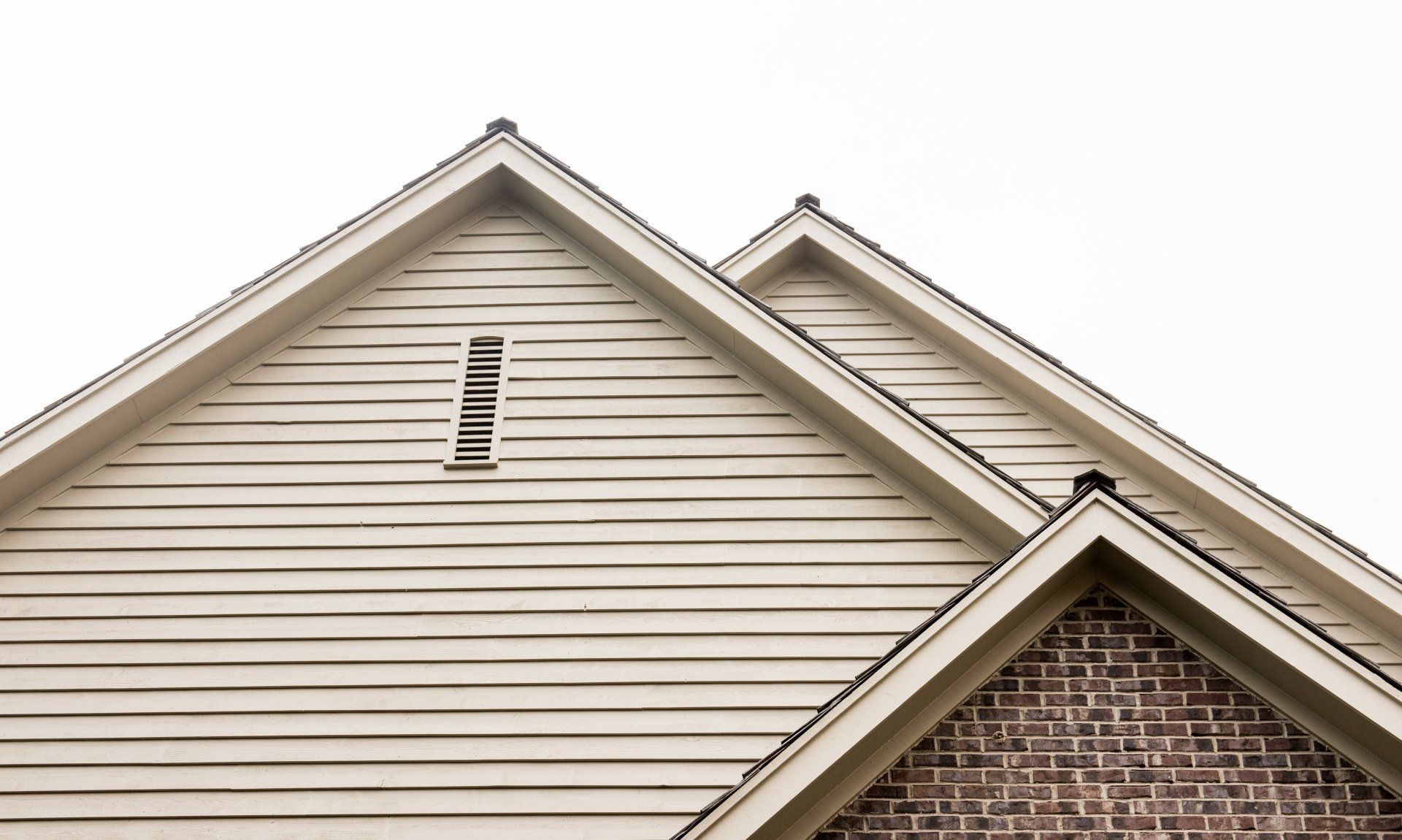 Side view of two gabled house roofs with beige siding and red brick trim against a white sky
