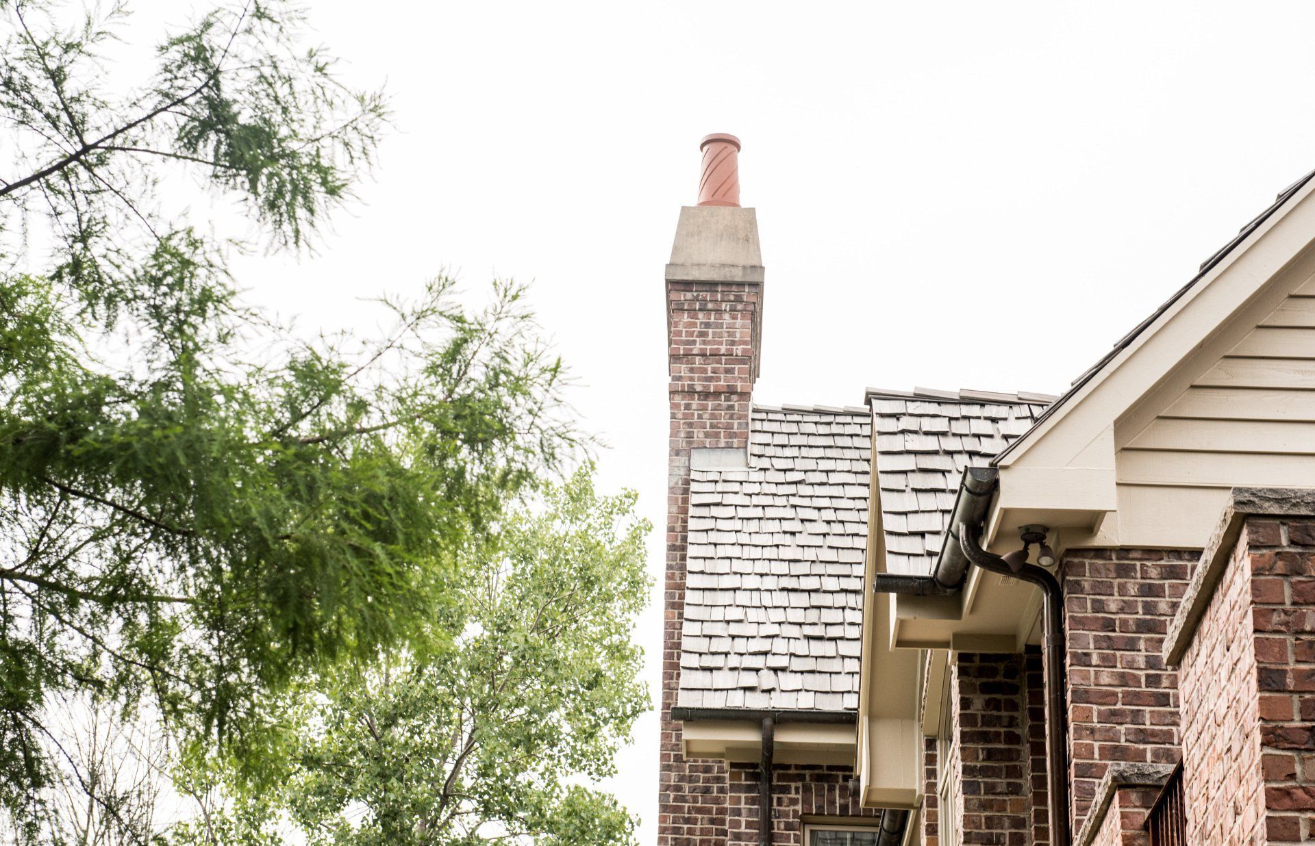 Brick house roof and chimney beside a leafy tree against a bright sky