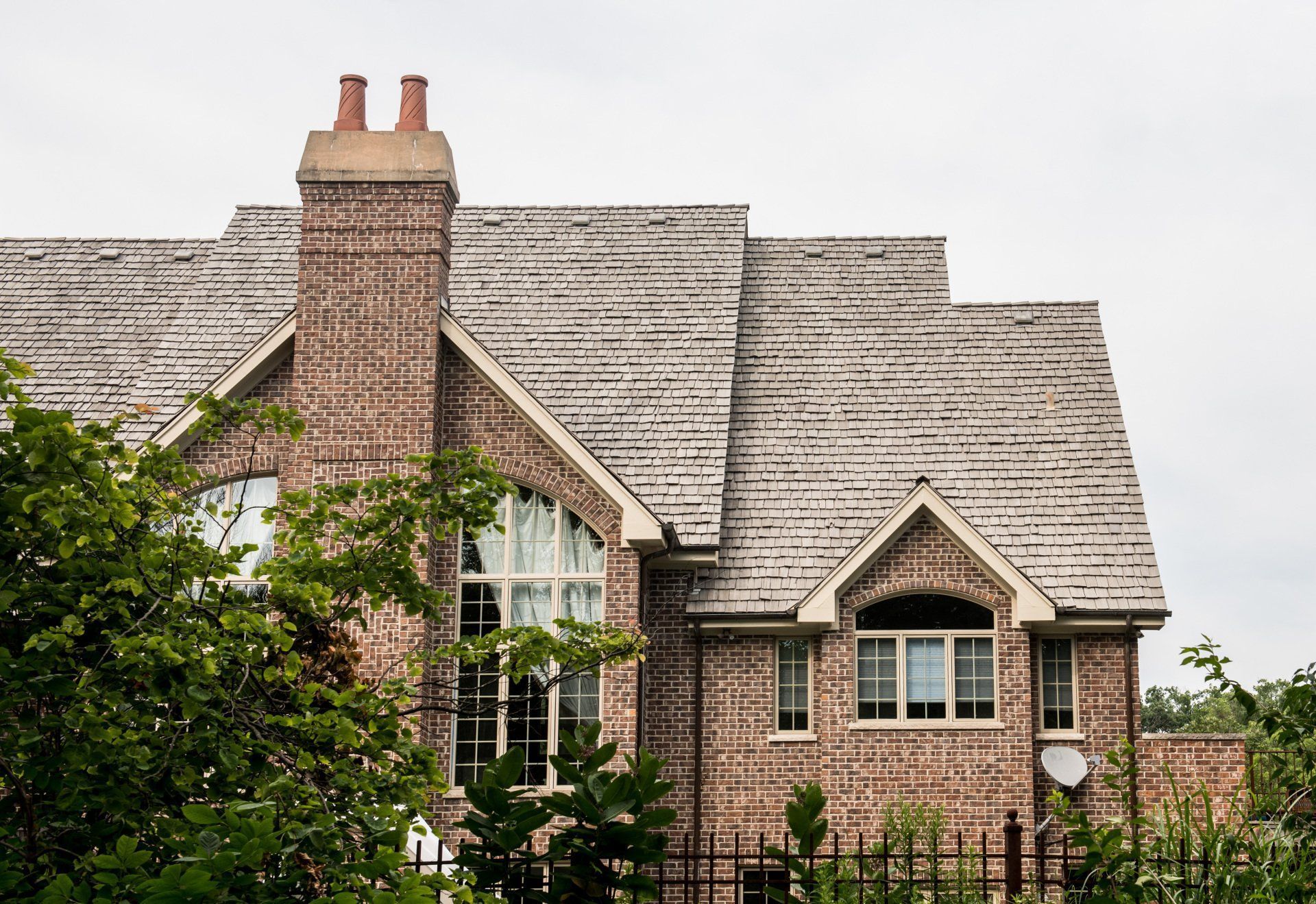 Brick house with steep gray shingle roof, large chimney, and green trees in front