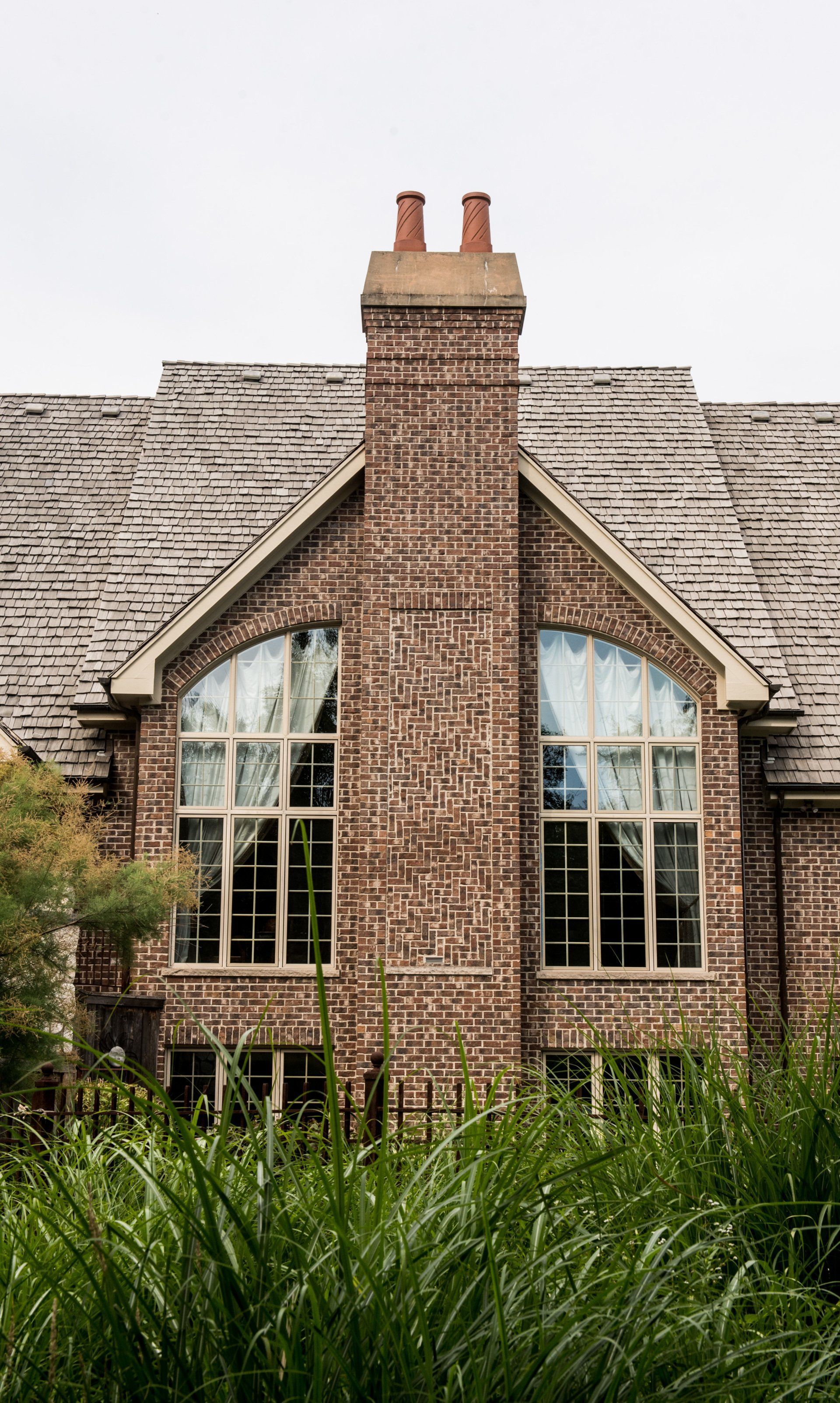 Brick house with a central chimney, arched windows, and tall grass in the foreground