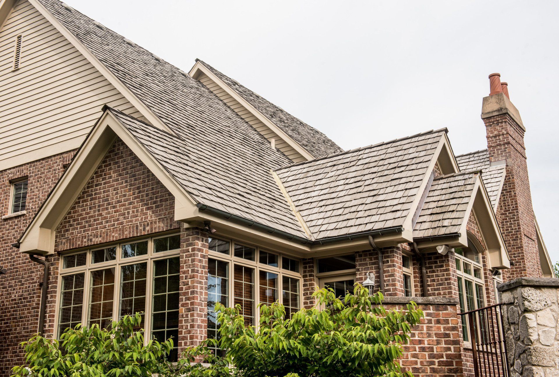 Brick house with steep shingled roof, large windows, and a chimney behind green shrubs