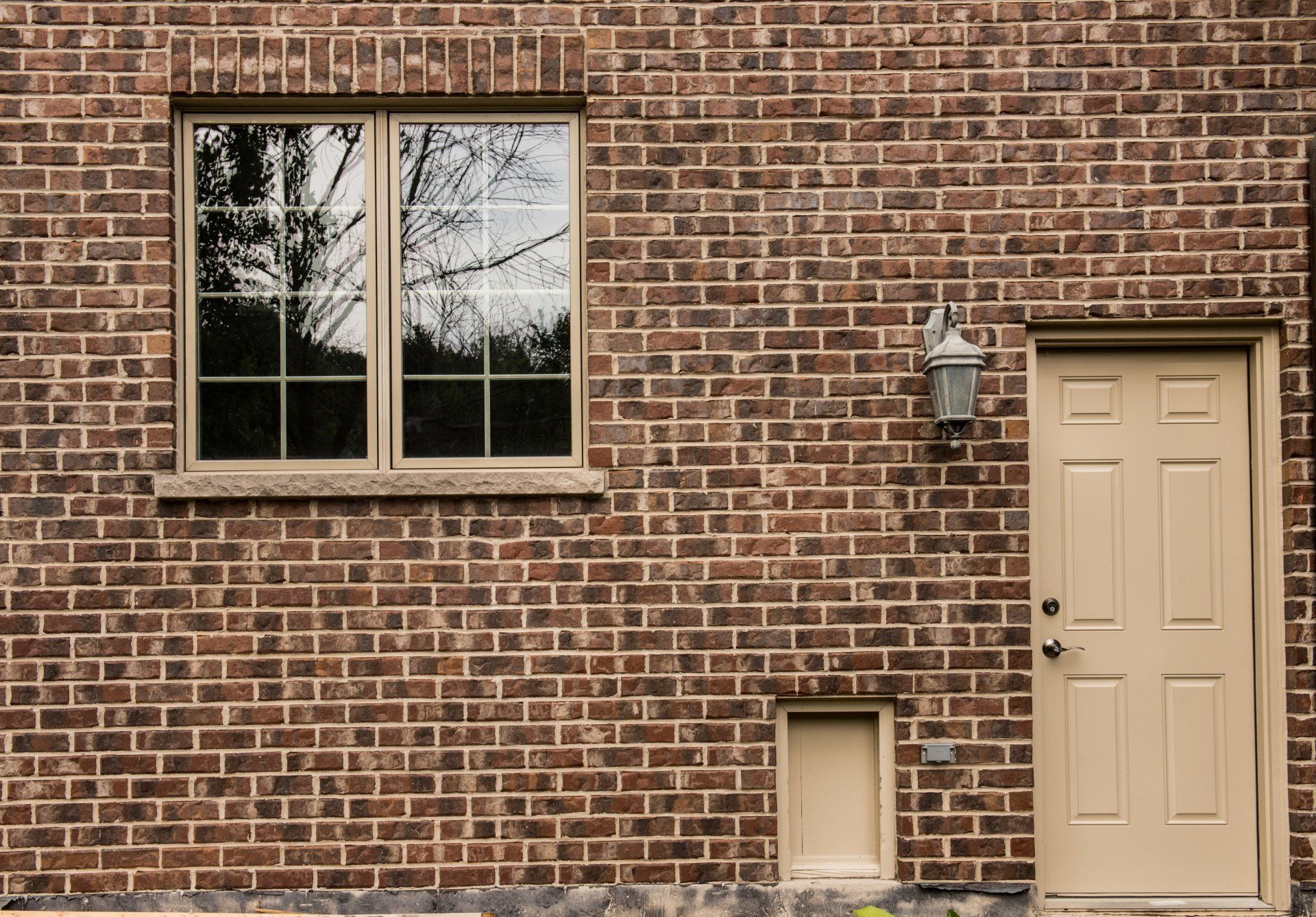 Brick building facade with a window, beige door, wall lantern, and small basement vent.