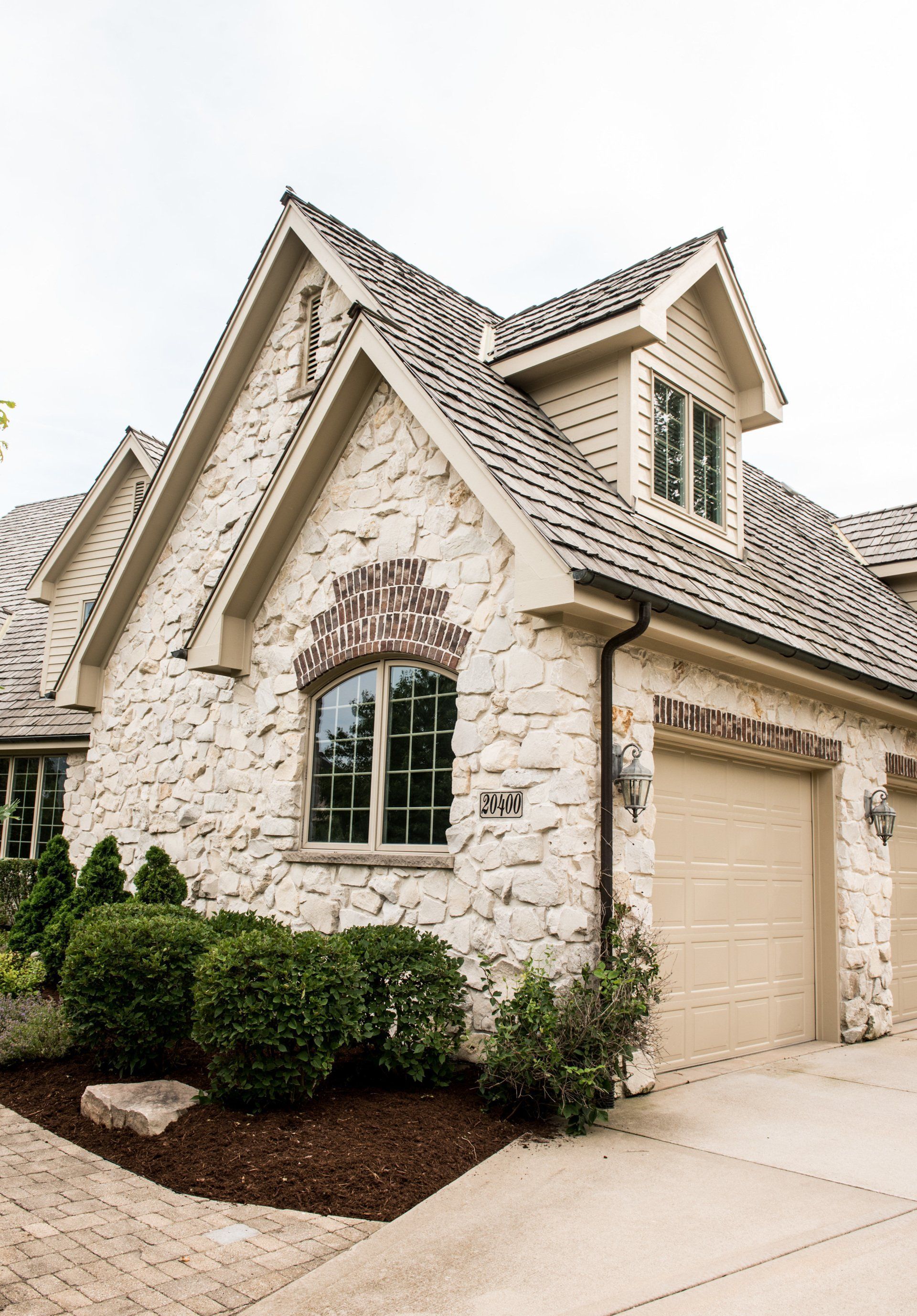 Stone house exterior with steep gables, arched window, beige garage, and trimmed shrubs along the driveway