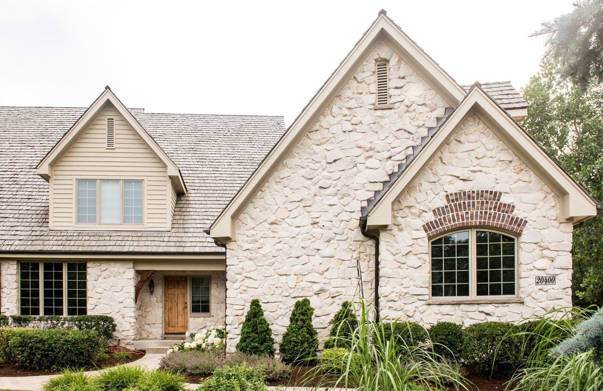 Stone house with beige siding, peaked rooflines, and a front garden of shrubs and greenery