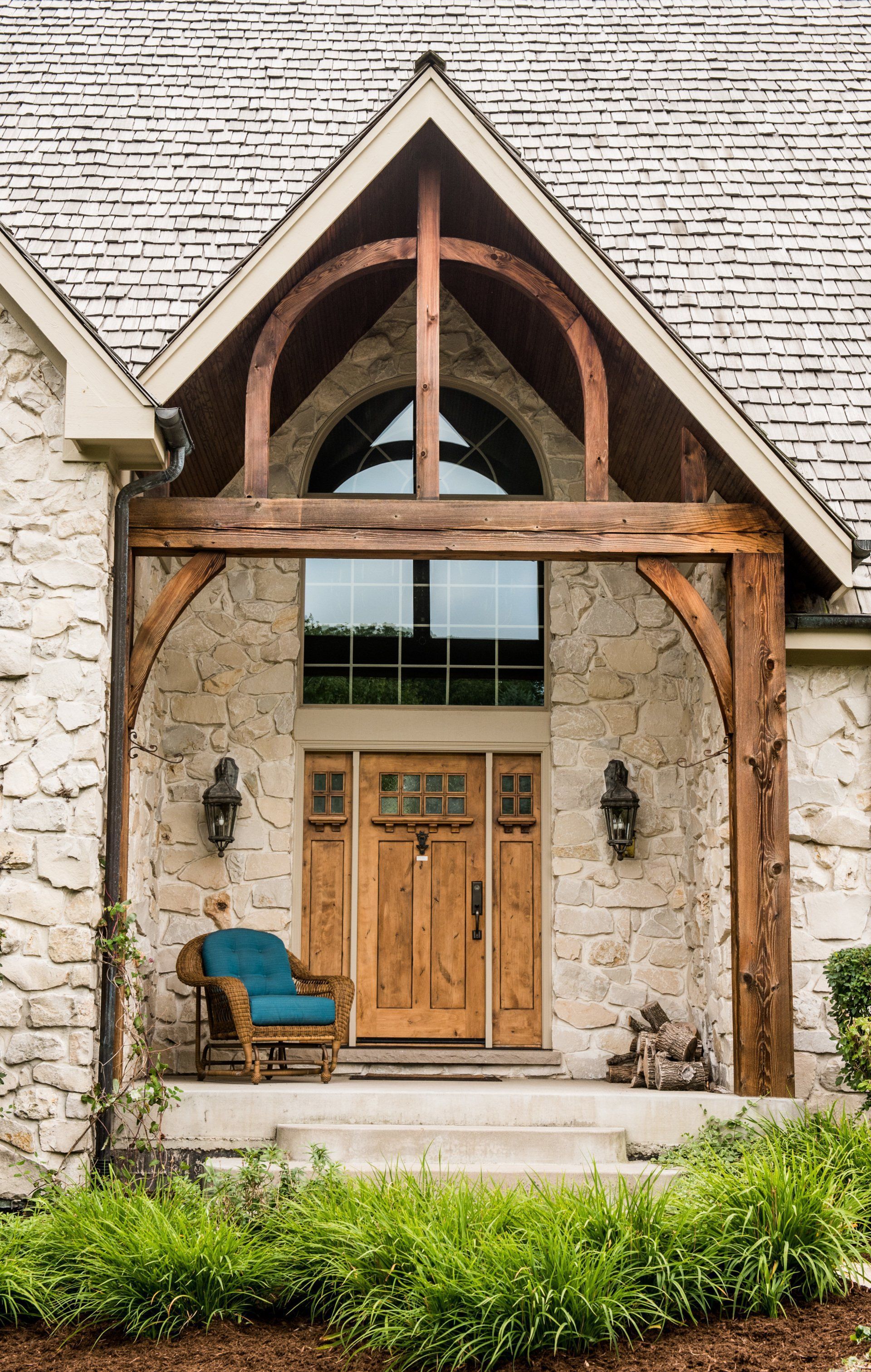 Stone house entry with rustic timber awning, arched window, wooden double doors, and teal chair on the porch