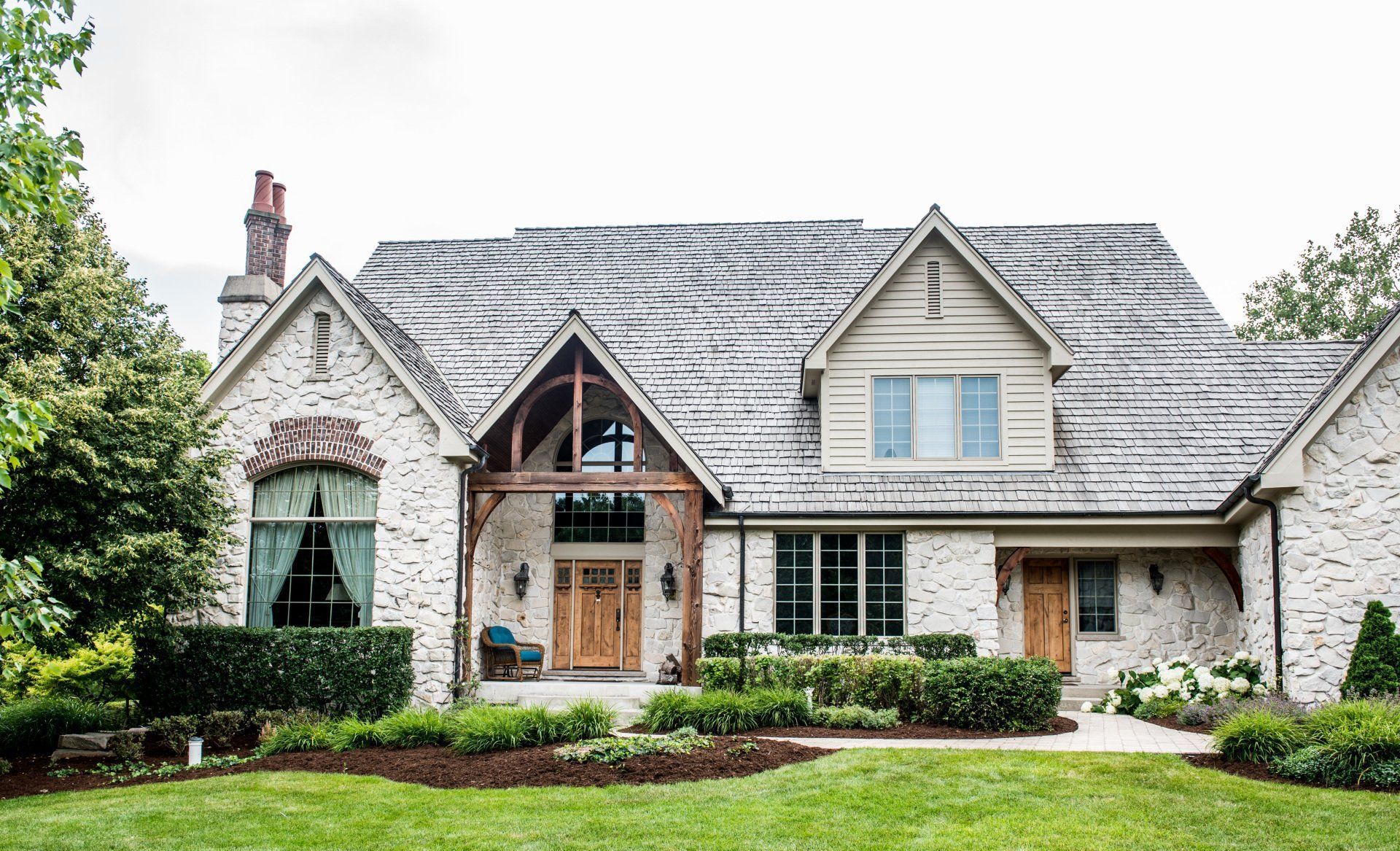 Stone house with gray shingle roof, dormer windows, and arched entry, surrounded by trees and a green lawn