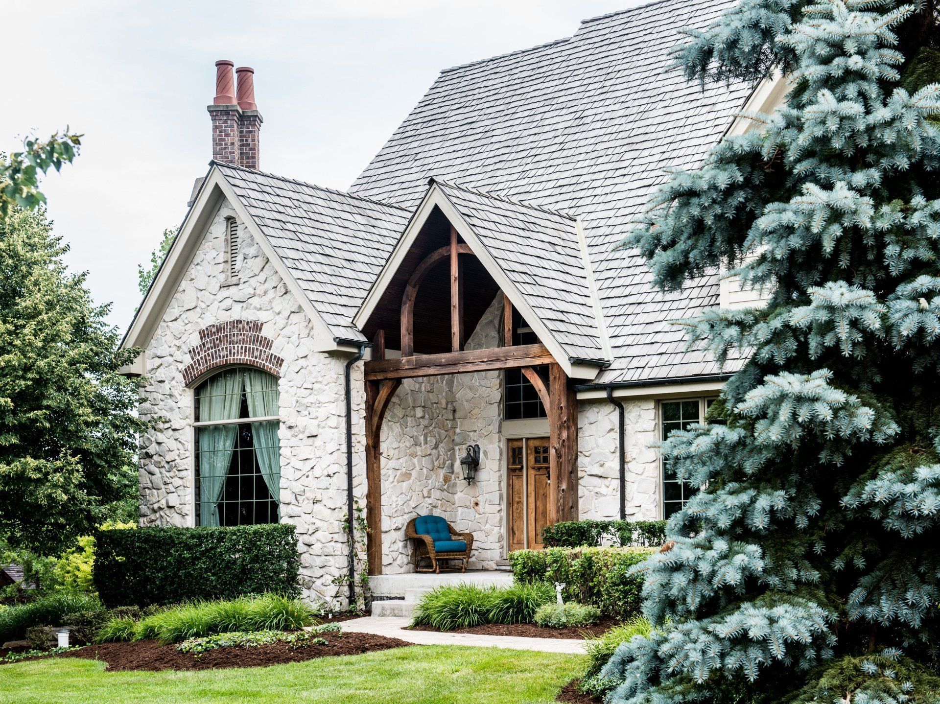 Stone cottage with steep roof, arched porch, and lush green landscaping, framed by evergreen trees