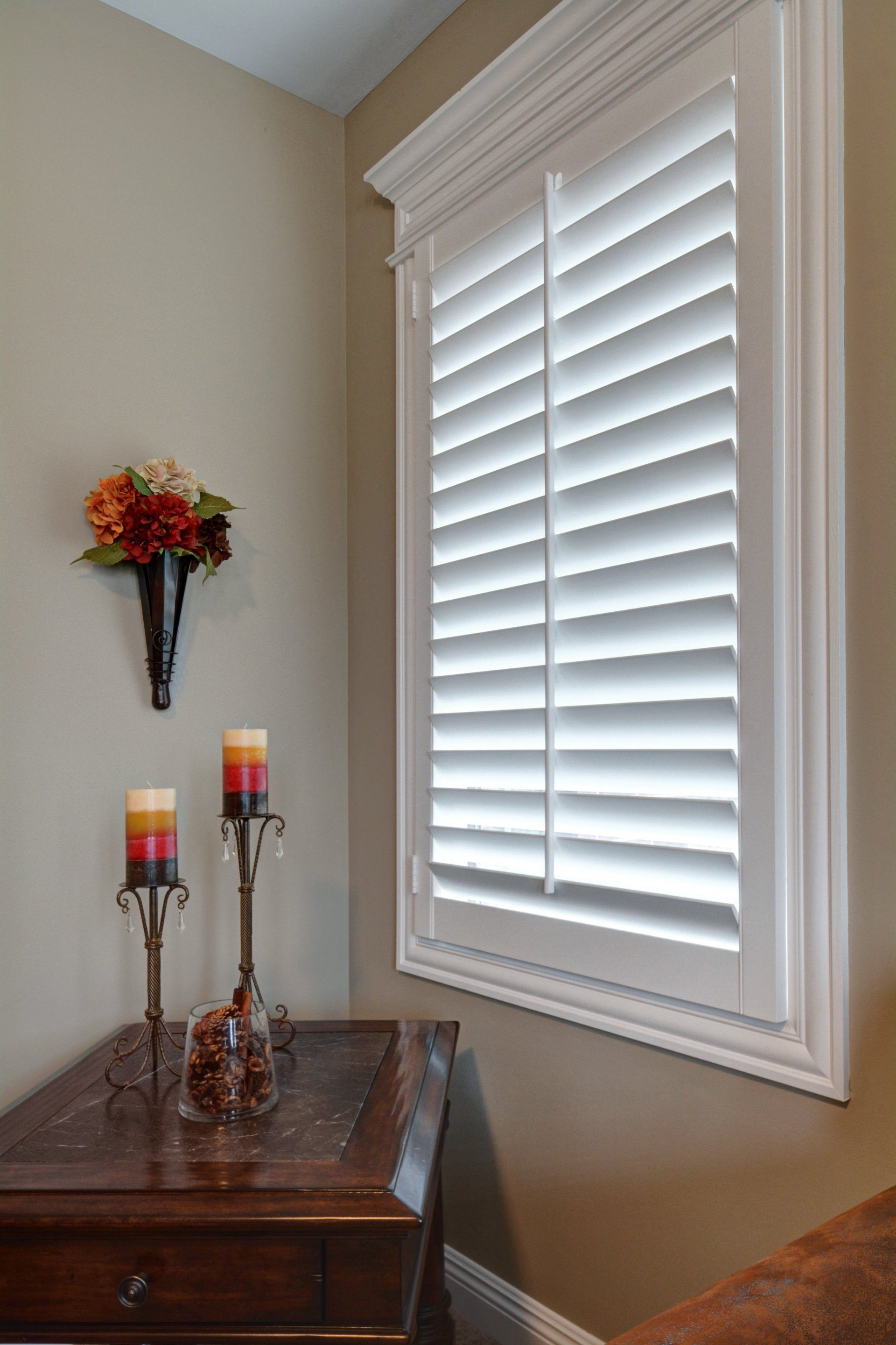 Corner of a room with white window shutters, a wooden table, and decorative candles and flowers.