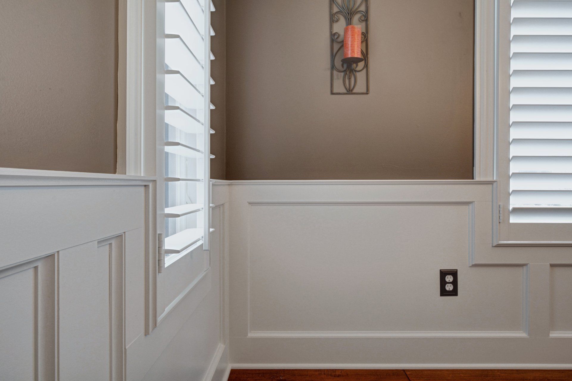 Empty hallway with beige walls, white wainscoting, and a wall sconce between shuttered windows.
