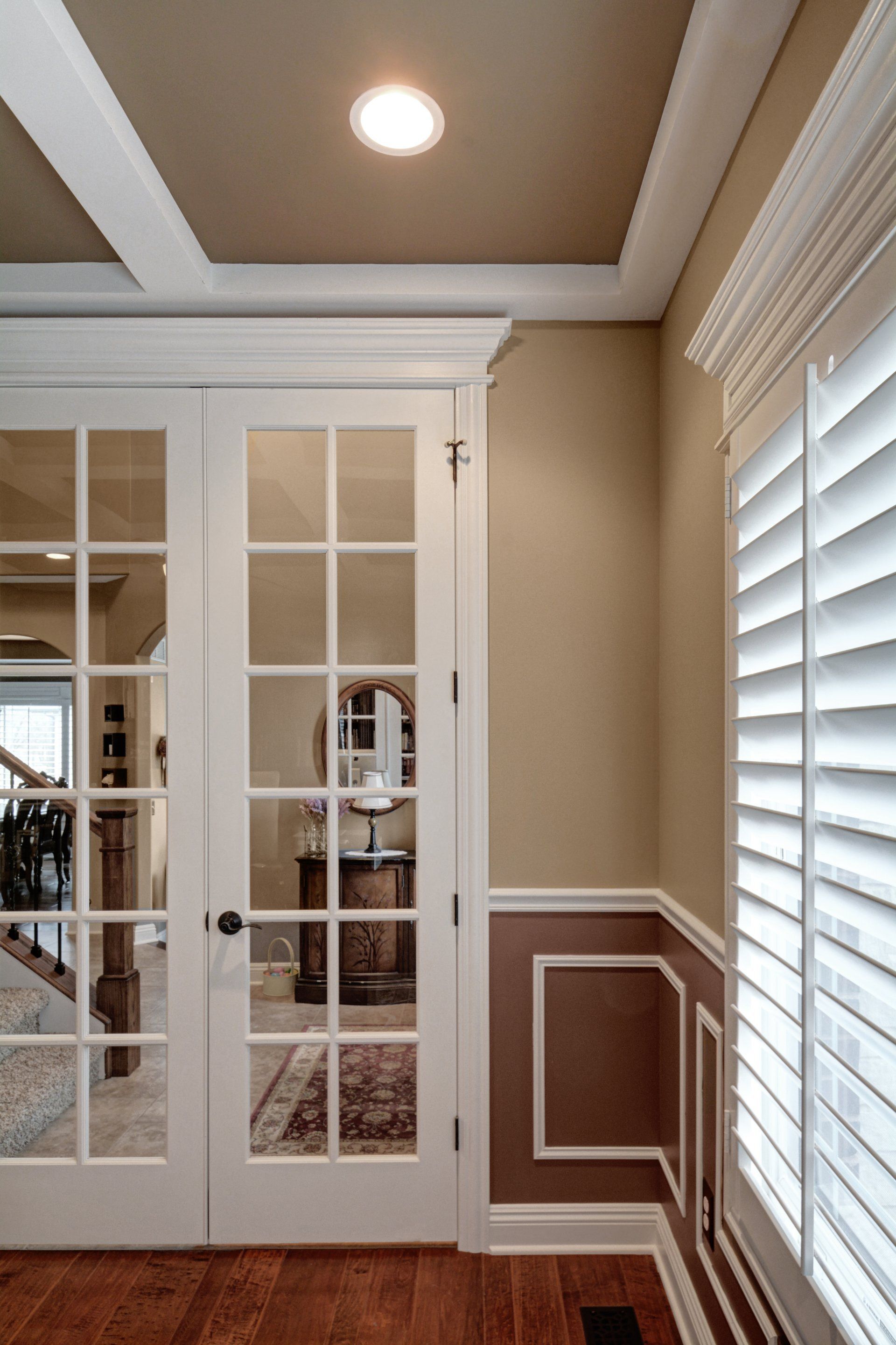 Elegant hallway with white French doors, beige walls, crown molding, and a window with blinds.