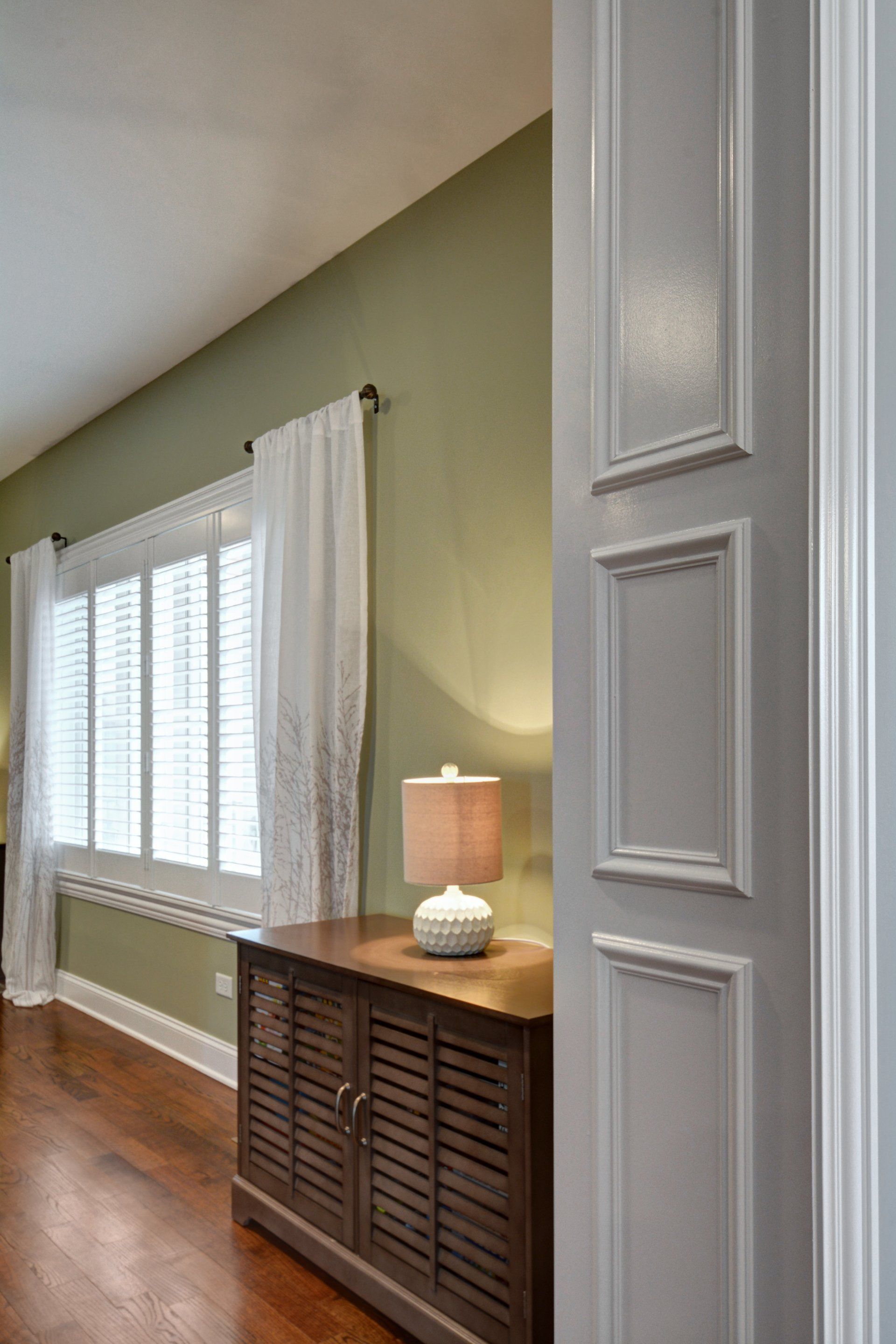 Bright hallway with green walls, white window curtains, and a wood cabinet topped by a lit lamp.