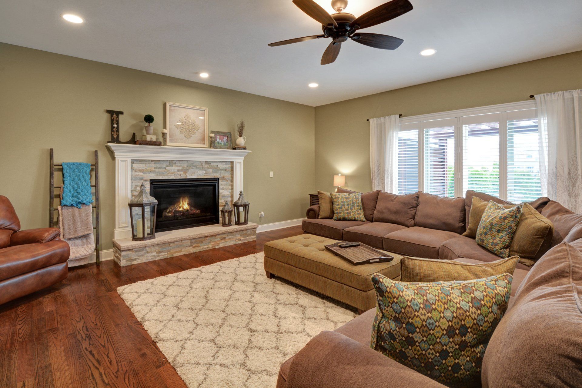 Living room with stone fireplace, sectional sofa, ceiling fan, and sunlit window shutters