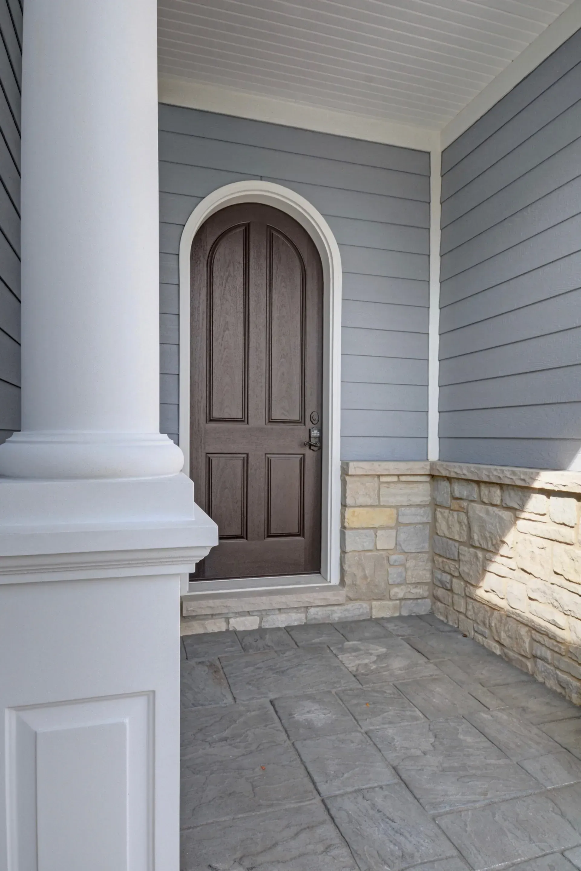 Front porch with a brown arched door, white column, gray siding, and stone-trimmed wall.