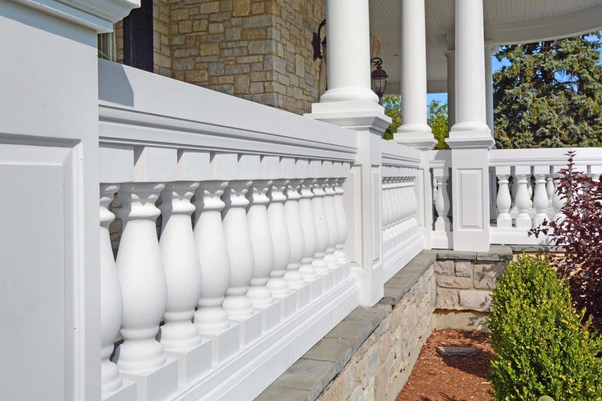 White balustrade and porch columns beside a stone house with garden plants and a brick walkway.