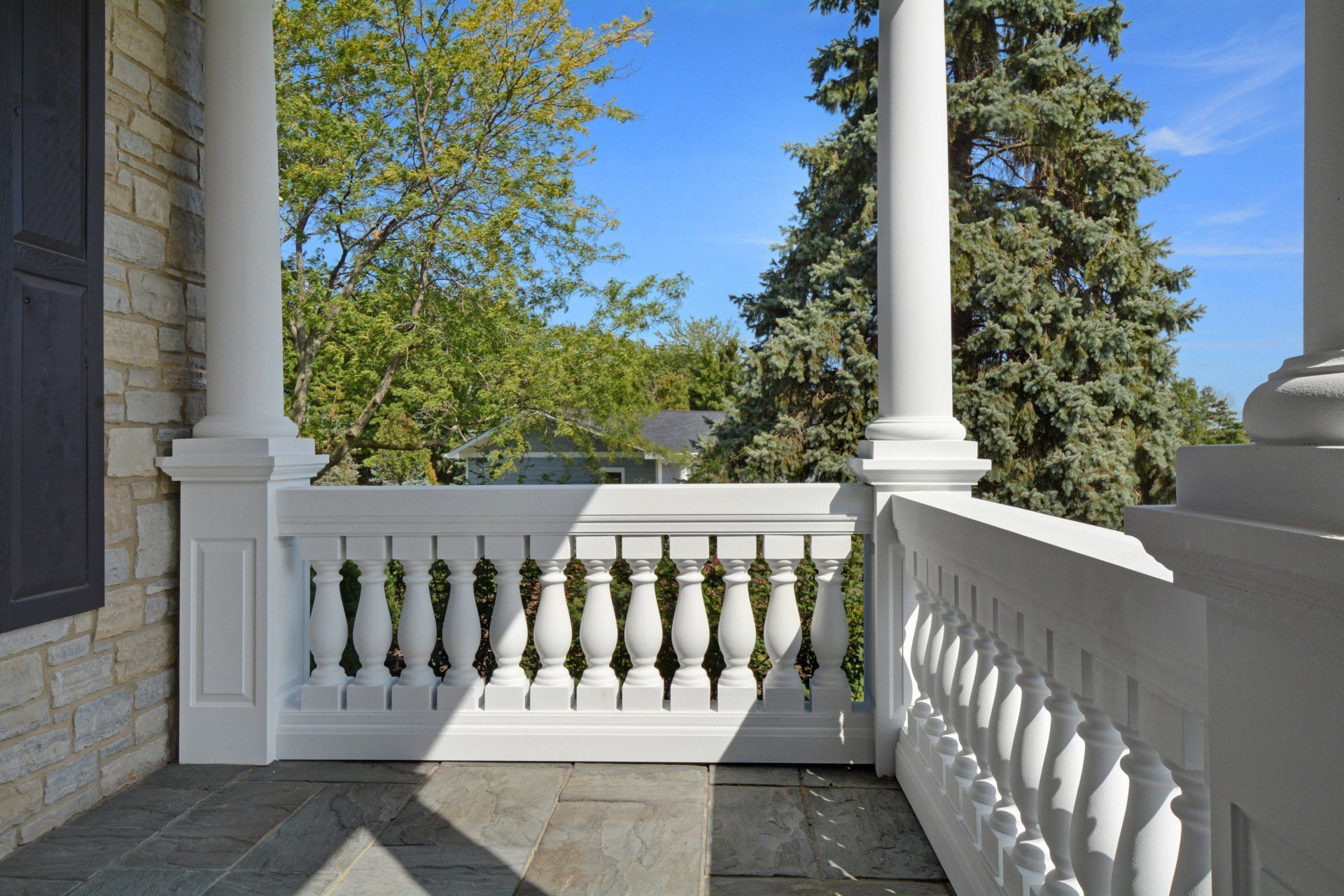 Sunlit porch with white balustrade and columns overlooking leafy trees and blue sky