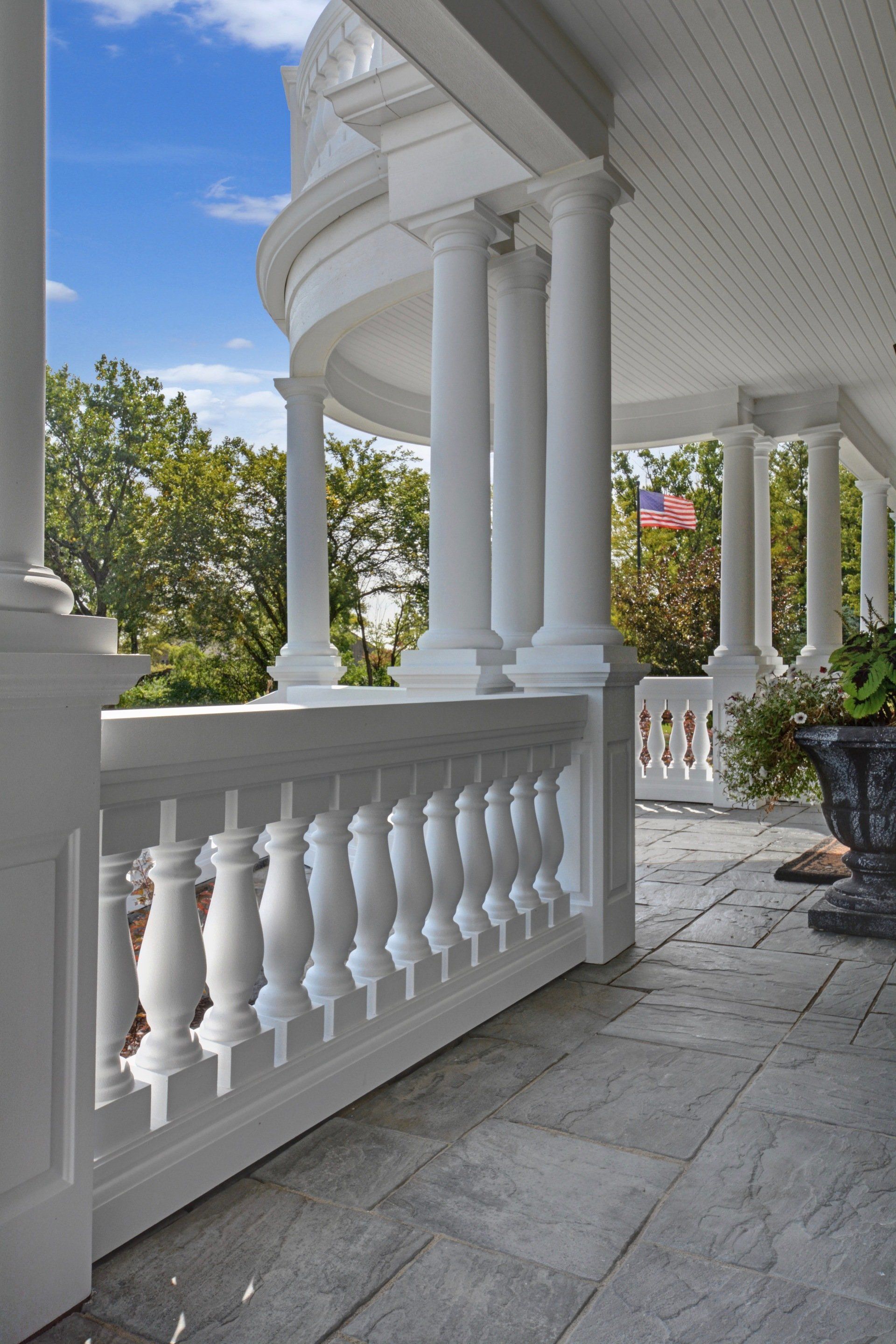 White-columned porch with balustrade, stone floor, and greenery under a bright blue sky
